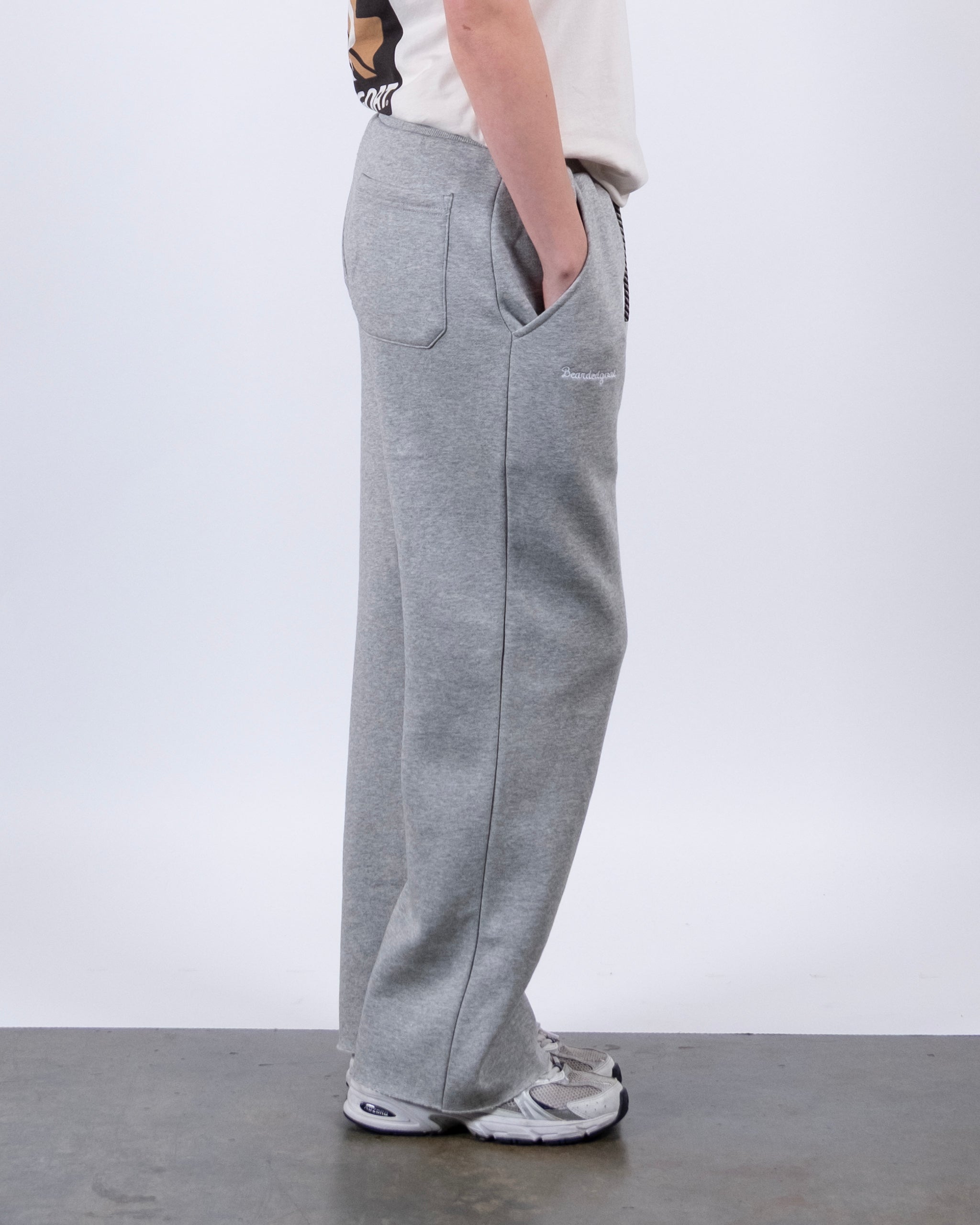 A person stands in side view wearing a white t-shirt, white sneakers, and beardedgoat Women’s Overnight Sweatpant made from heavyweight cotton fleece with pockets. The background is plain and light-colored.