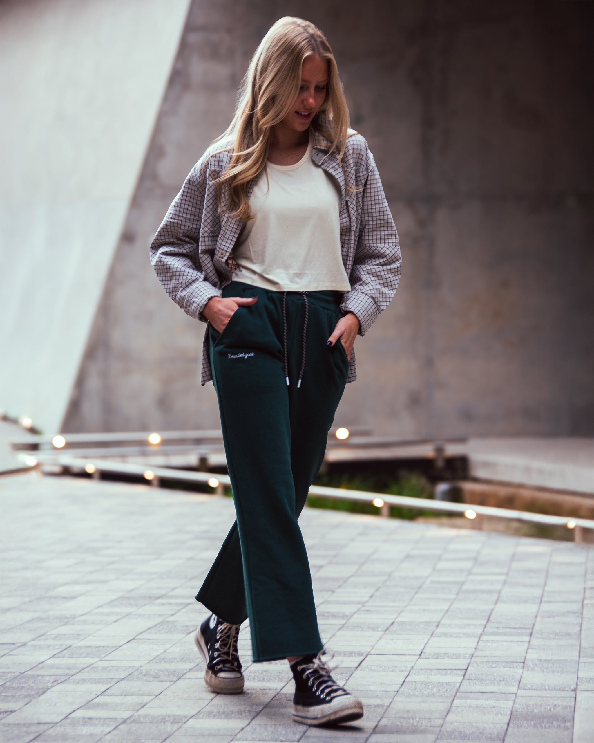 A woman with long blonde hair walks outside on a tiled path, smiling in a casual look: light shirt, plaid jacket, beardedgoat Women’s Overnight Sweatpant made from heavyweight cotton fleece, and black Converse sneakers.