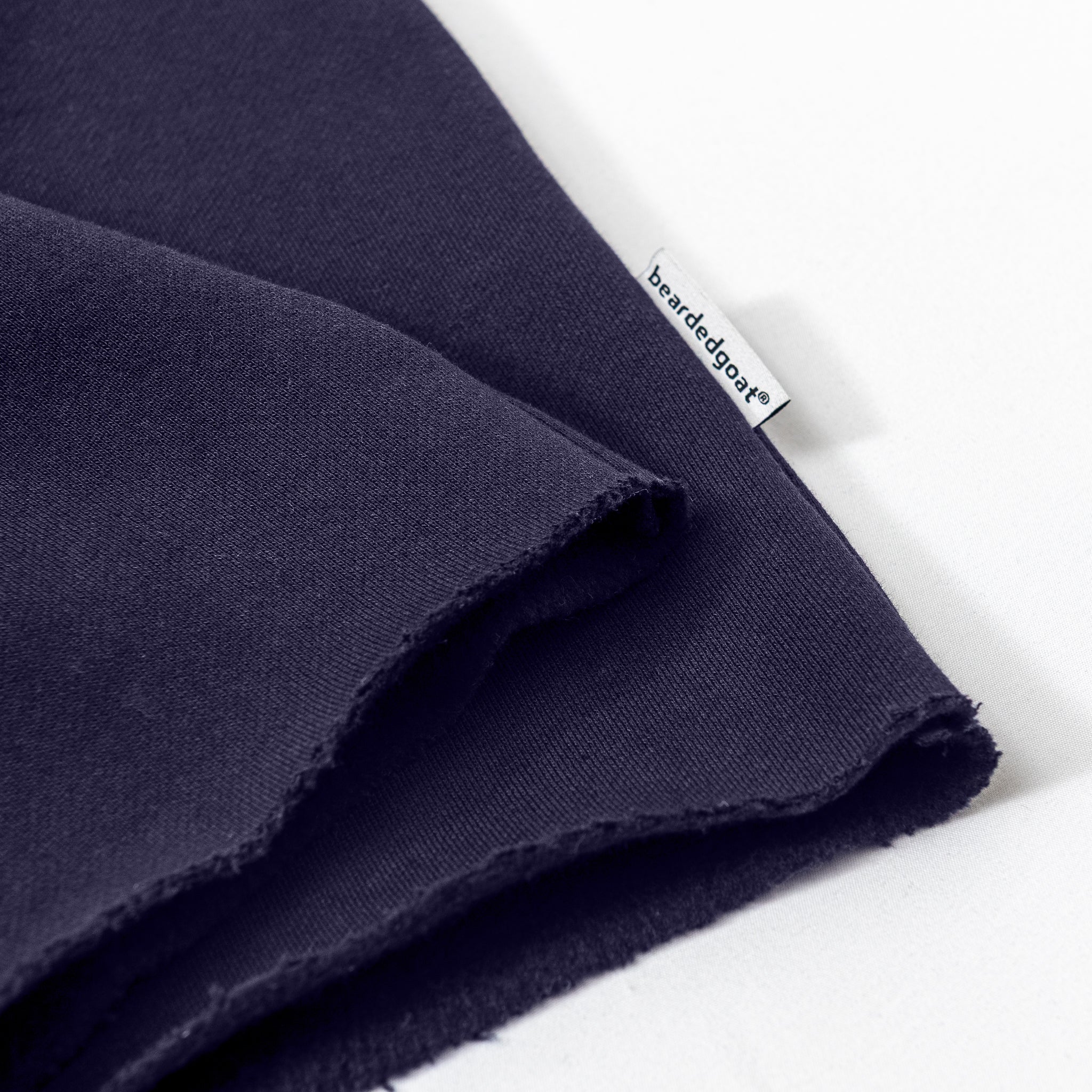 A close-up of heavyweight dark navy cotton fleece from the beardedgoat Women’s Overnight Sweatpant, folded with raw edges and a small white beardedgoat label sewn into the seam, shown on a white surface—ideal for comfortable loungewear.