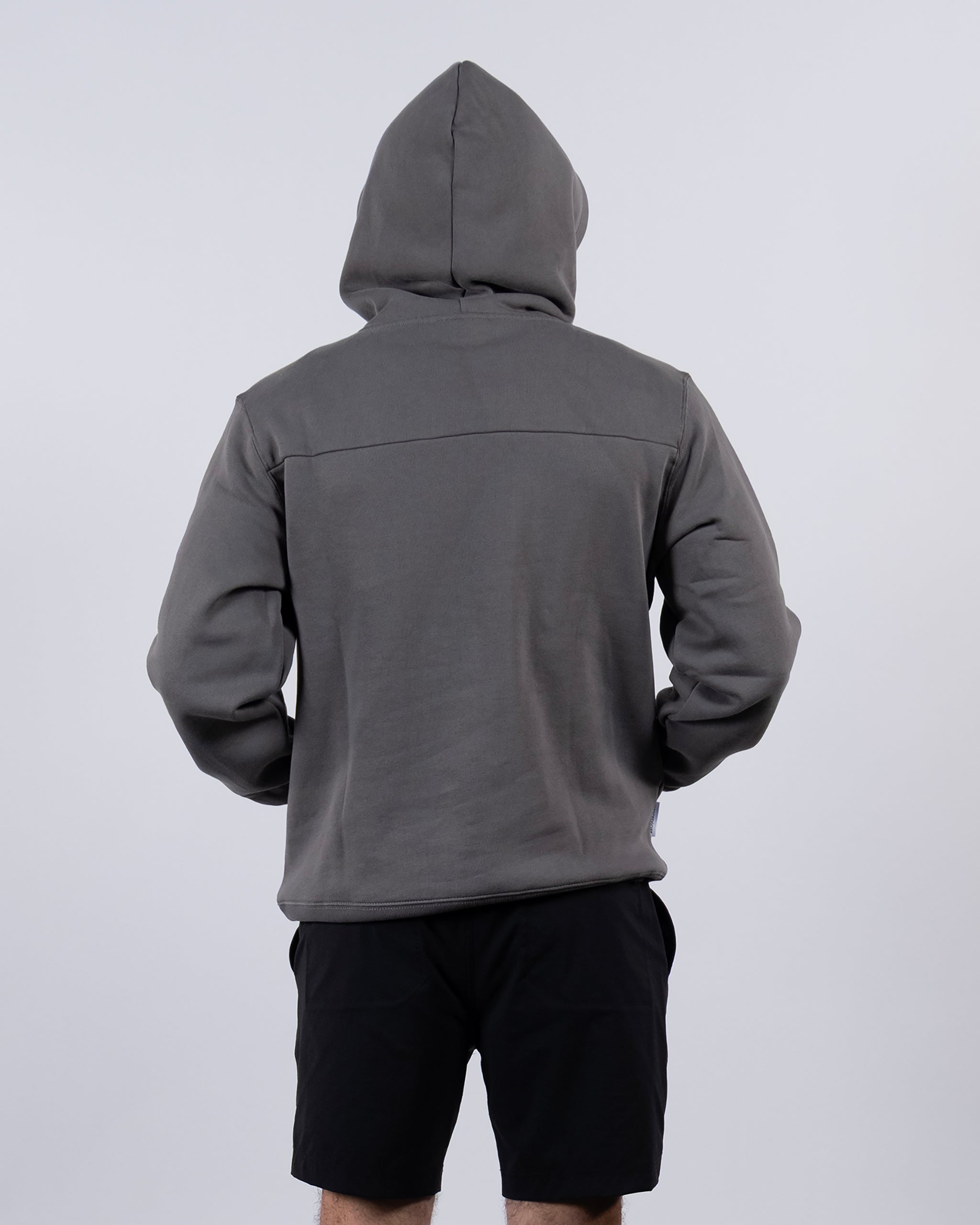 A person in a gray beardedgoat The VOID™ Hoodie, crafted from heavyweight cotton fleece with the hood up, and black shorts stands facing away against a plain light background.