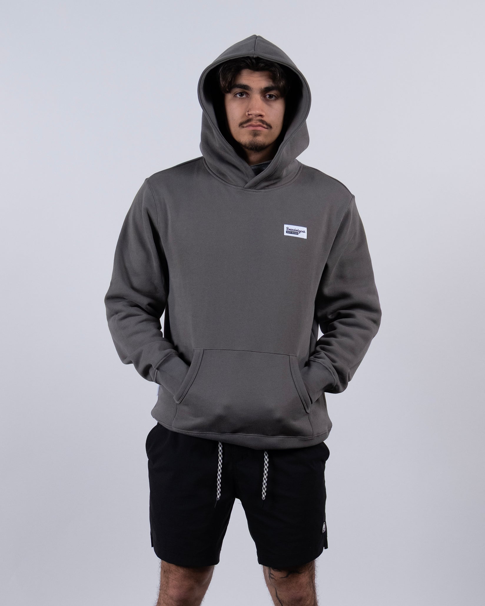 A person wearing the beardedgoat VOID™ Hoodie in gray stands against a plain, light background with the hood up, hands in the pocket, and looks directly at the camera.