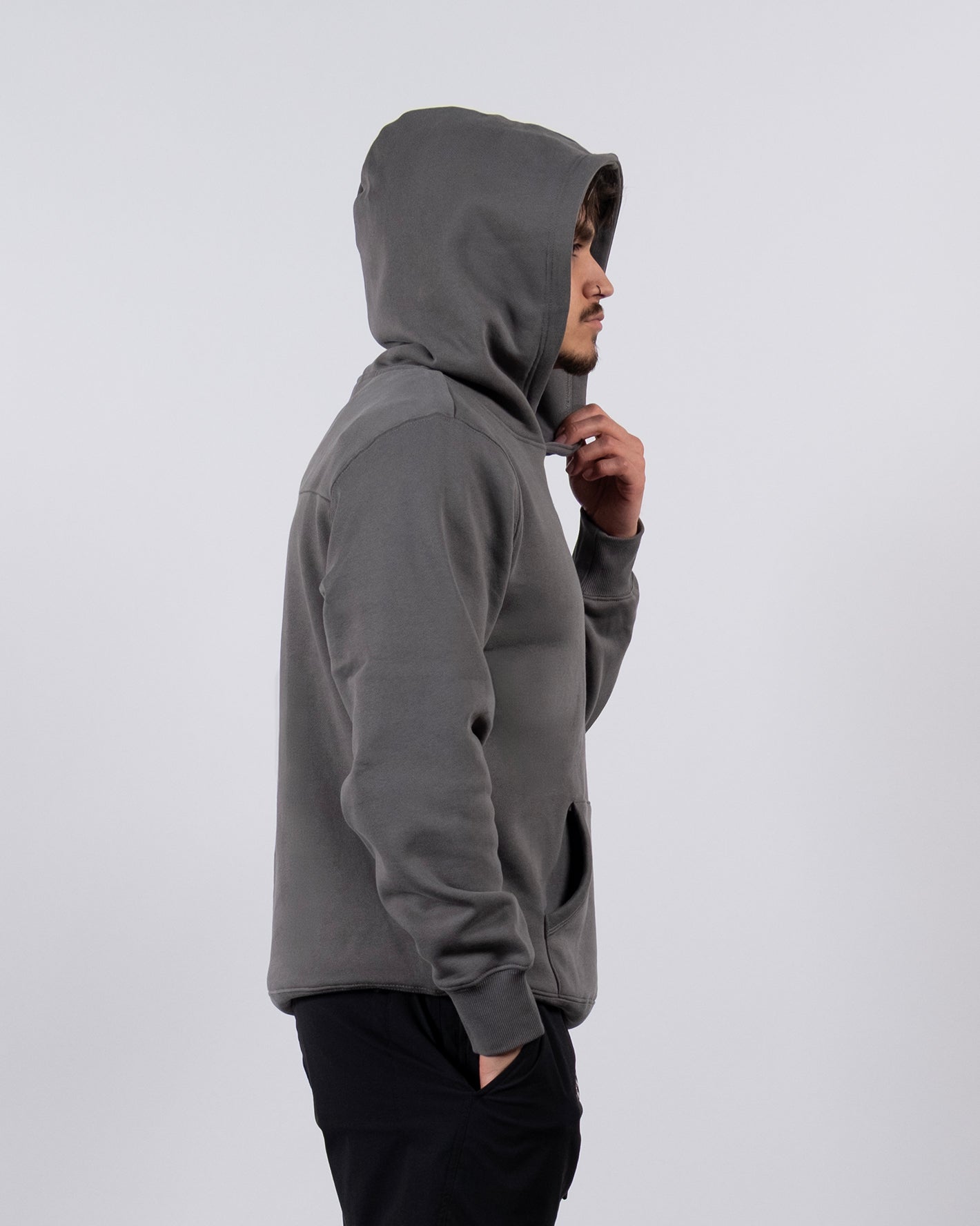 A person in profile stands against a plain light gray background, wearing the beardedgoat VOID™ Hoodie in gray (heavyweight cotton fleece, hood up) paired with black pants.