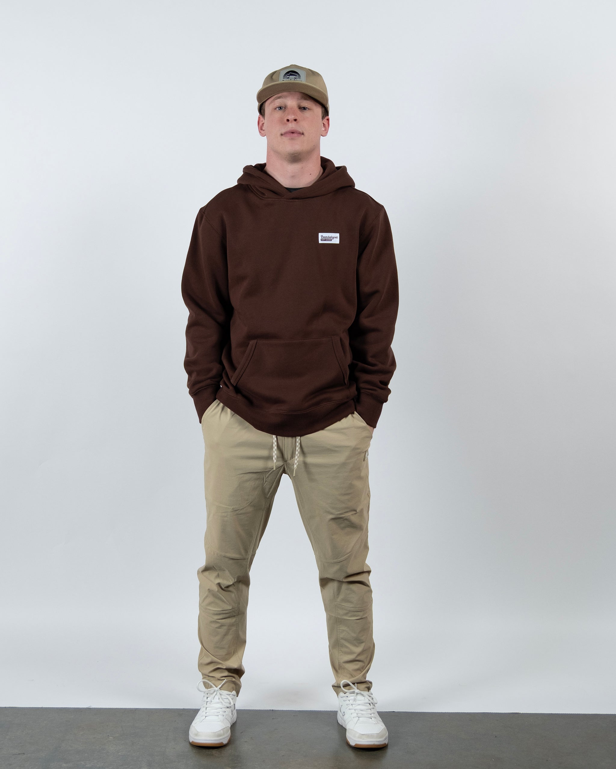 A person stands against a white background wearing The VOID™ Hoodie by beardedgoat in brown heavyweight cotton fleece, paired with khaki pants, white sneakers, and a beige cap. Hands are in the hoodie pockets as they face the camera neutrally.