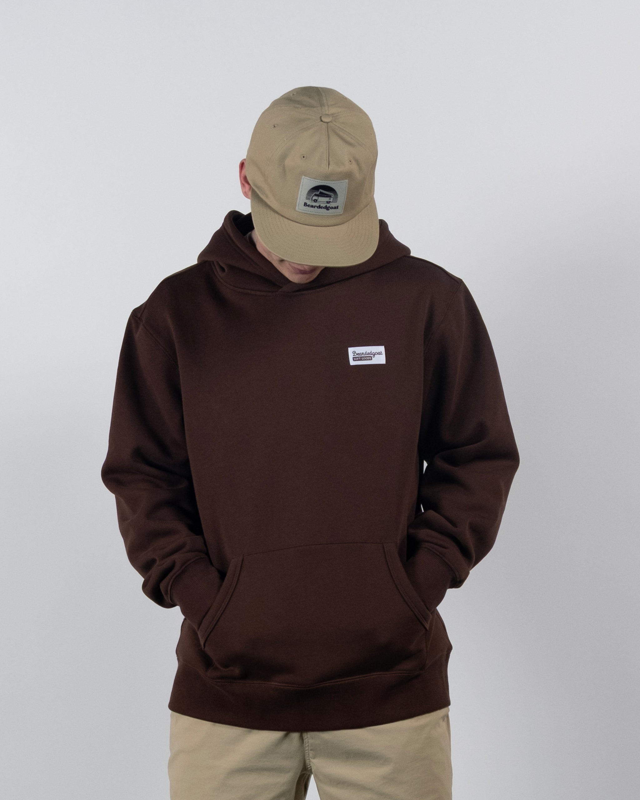 A person wearing The VOID™ Hoodie by beardedgoat—crafted from heavyweight cotton fleece in brown—with hands in the front pocket and a beige cap, looking down against a plain light background.