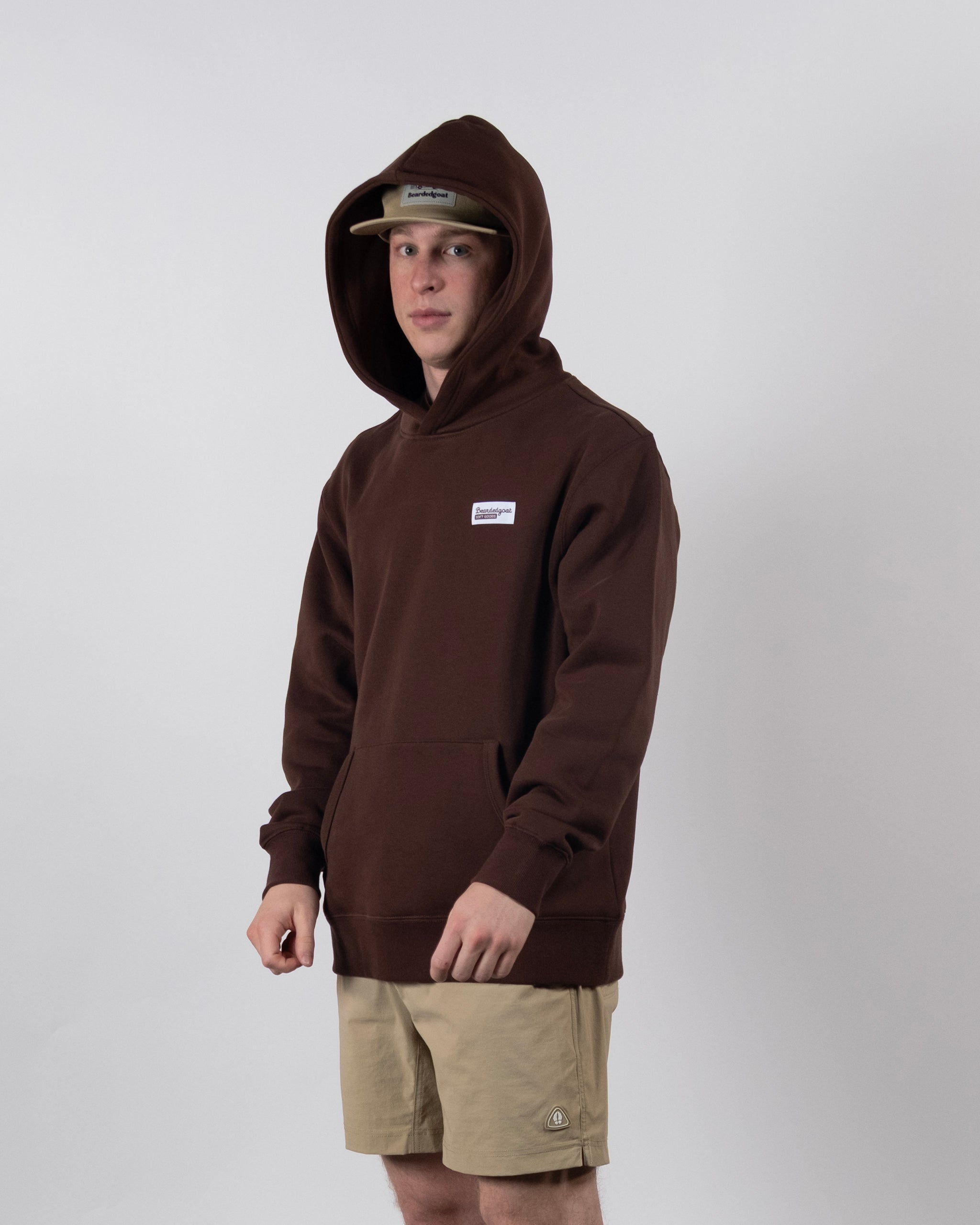 Wearing the beardedgoat The VOID™ Hoodie—a brown heavyweight cotton fleece—a person pairs it with tan shorts and a beige cap, standing against a plain light background and looking neutrally at the camera.