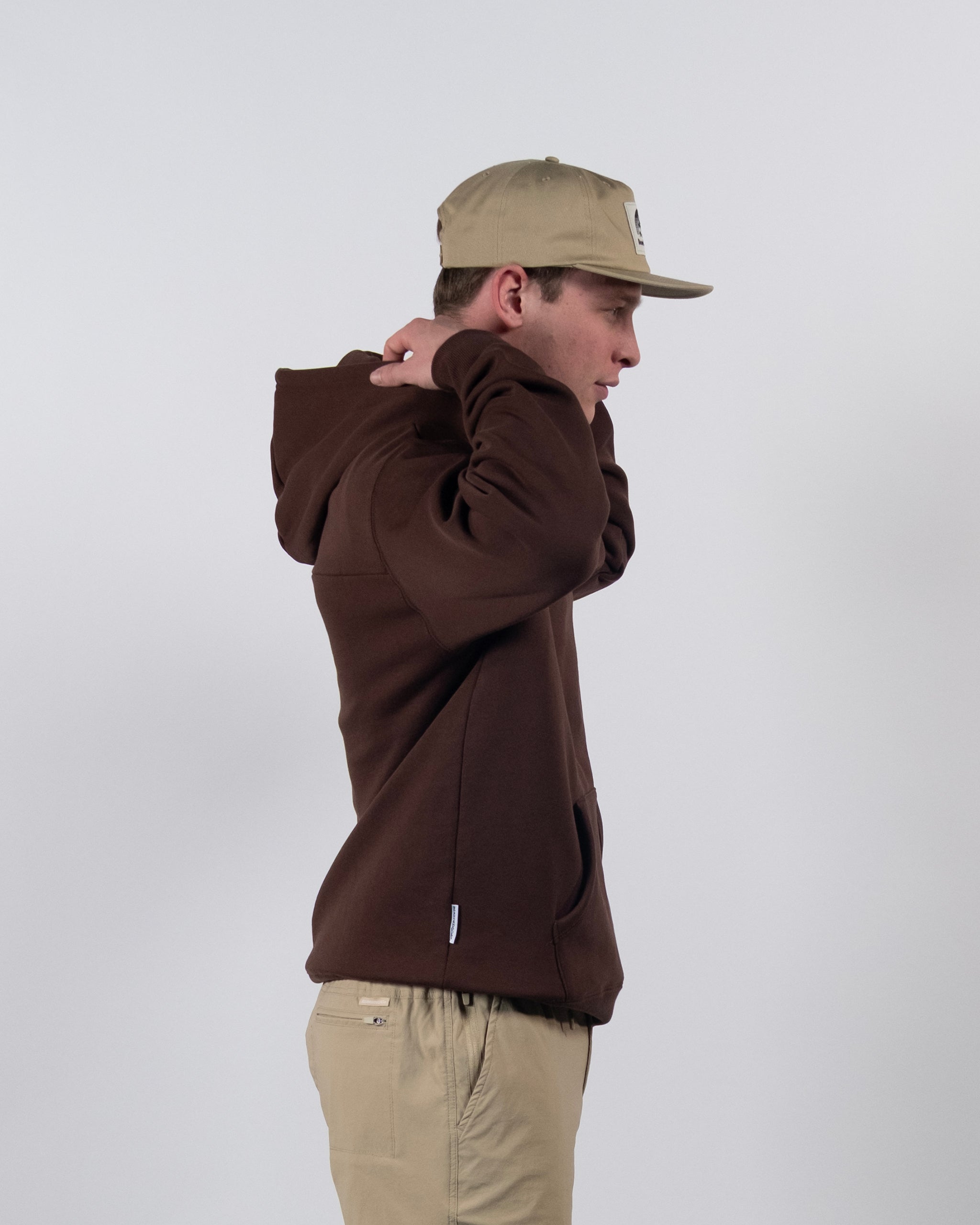 A person in a brown beardedgoat The VOID™ Hoodie made from heavyweight cotton fleece adjusts their hood. They stand sideways against a plain light background, wearing a beige cap and matching beige pants.