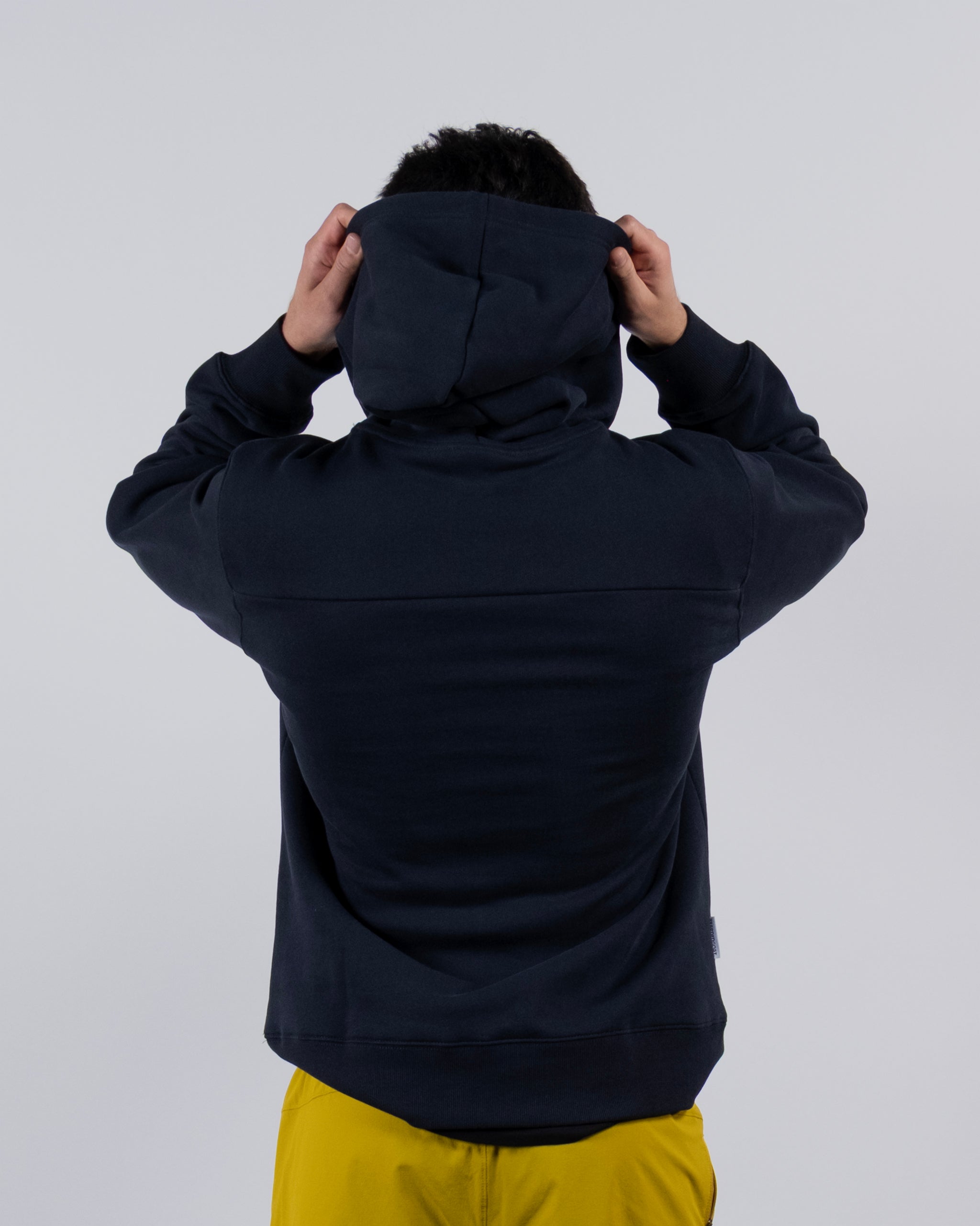 A person in a beardedgoat The VOID™ Hoodie and yellow pants stands with their back to the camera, holding up the hood with both hands against a plain light background.