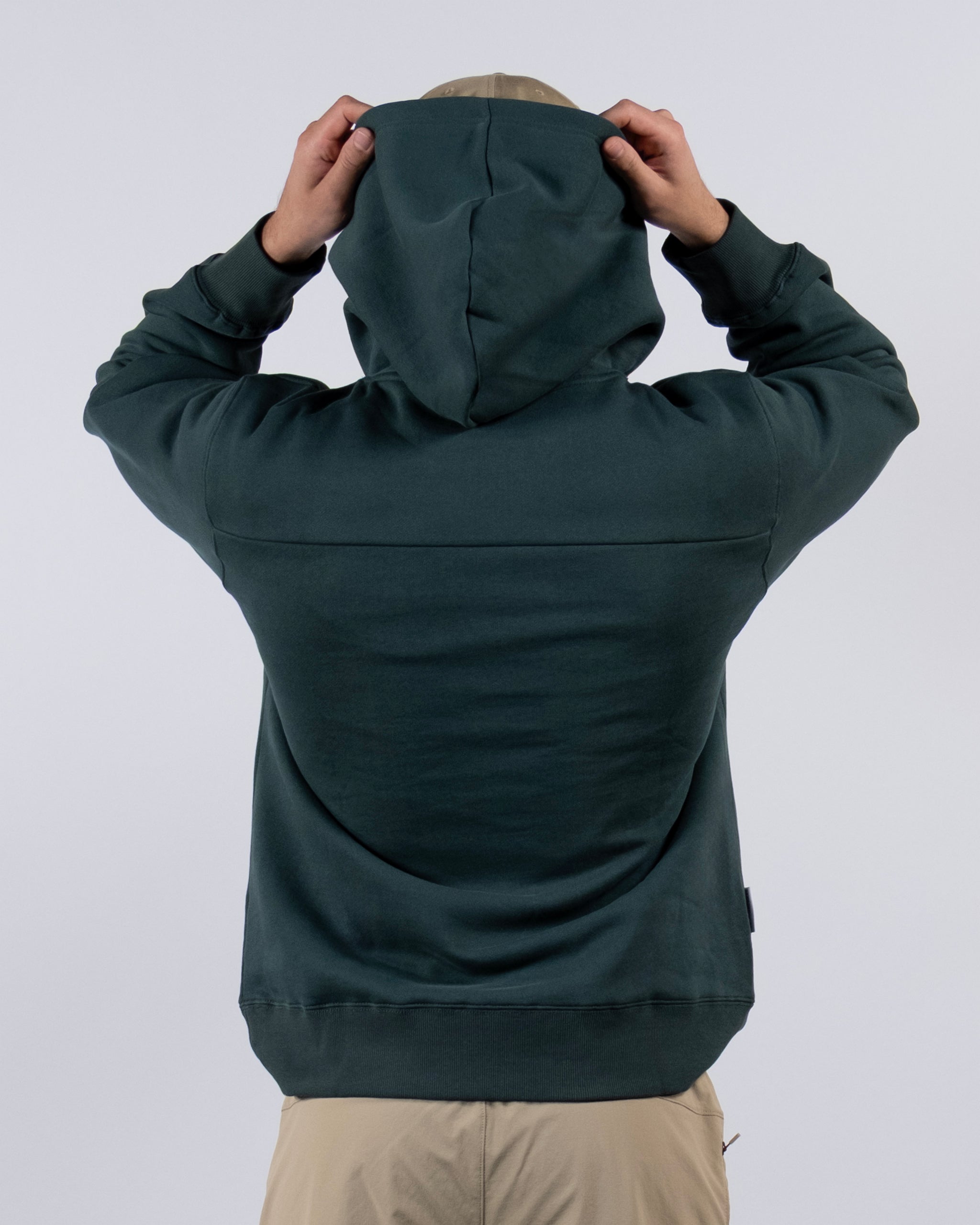 A person wearing the beardedgoat The VOID™ Hoodie, made from heavyweight cotton fleece, and beige pants faces away from the camera, lifting the hood with both hands against a plain light background.