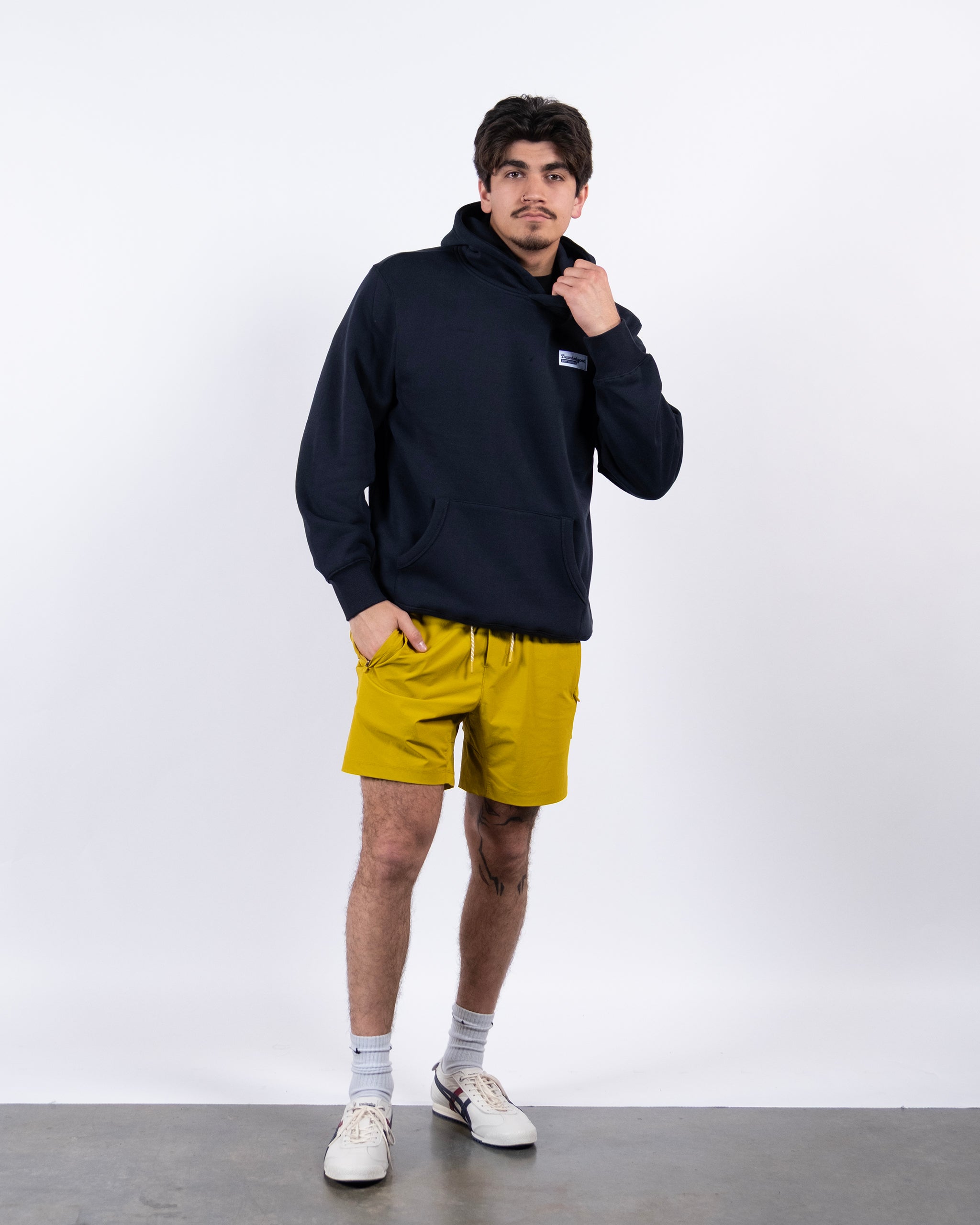 A man stands against a plain white background wearing the beardedgoat VOID™ Hoodie, yellow shorts, white socks, and white sneakers. He raises one hand to his collar and looks slightly to the side.