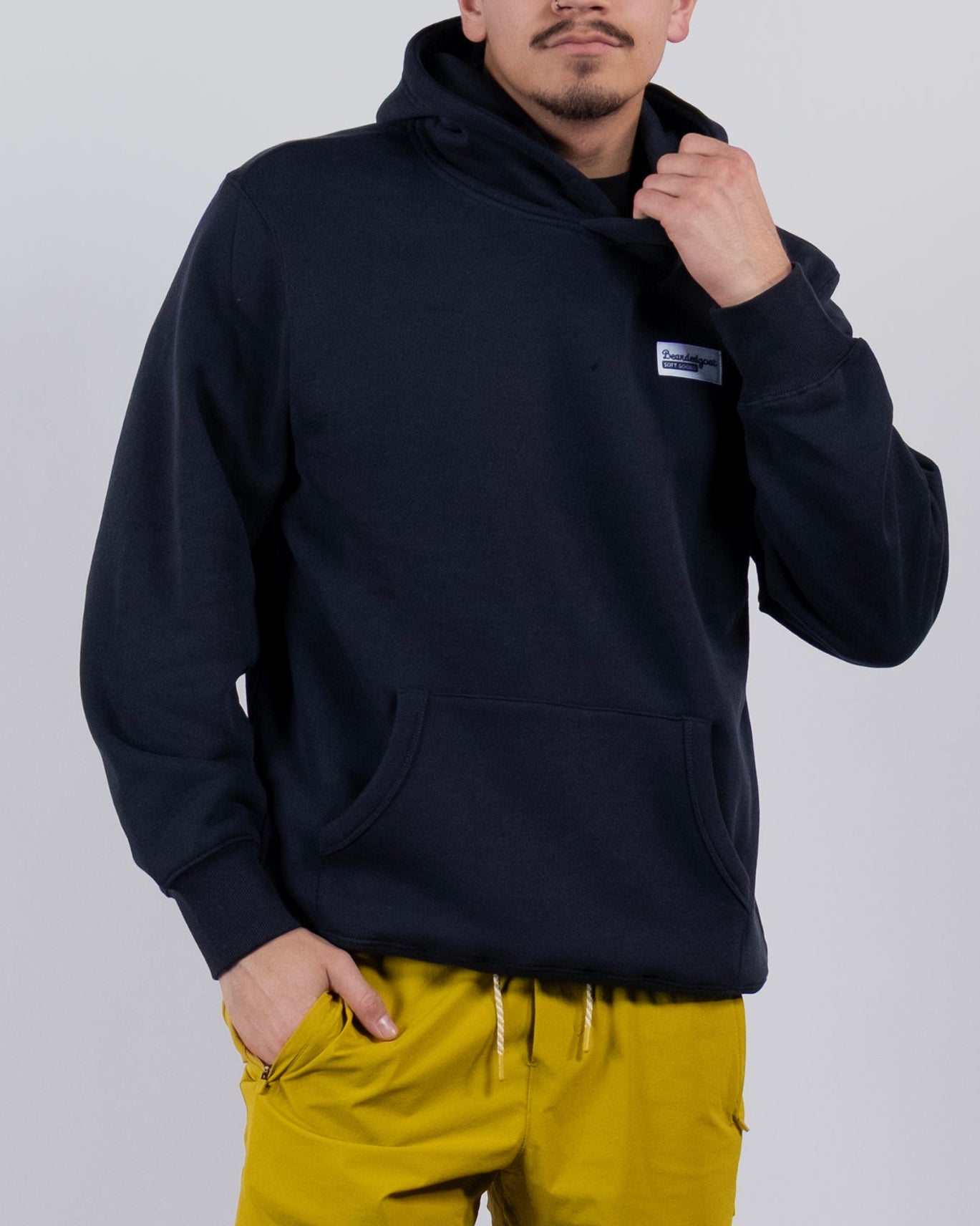 A man poses confidently against a plain background, wearing the best-selling beardedgoat The VOID™ Hoodie and bright yellow shorts, one hand in his pocket and the other adjusting his hoodie as he looks at the camera.