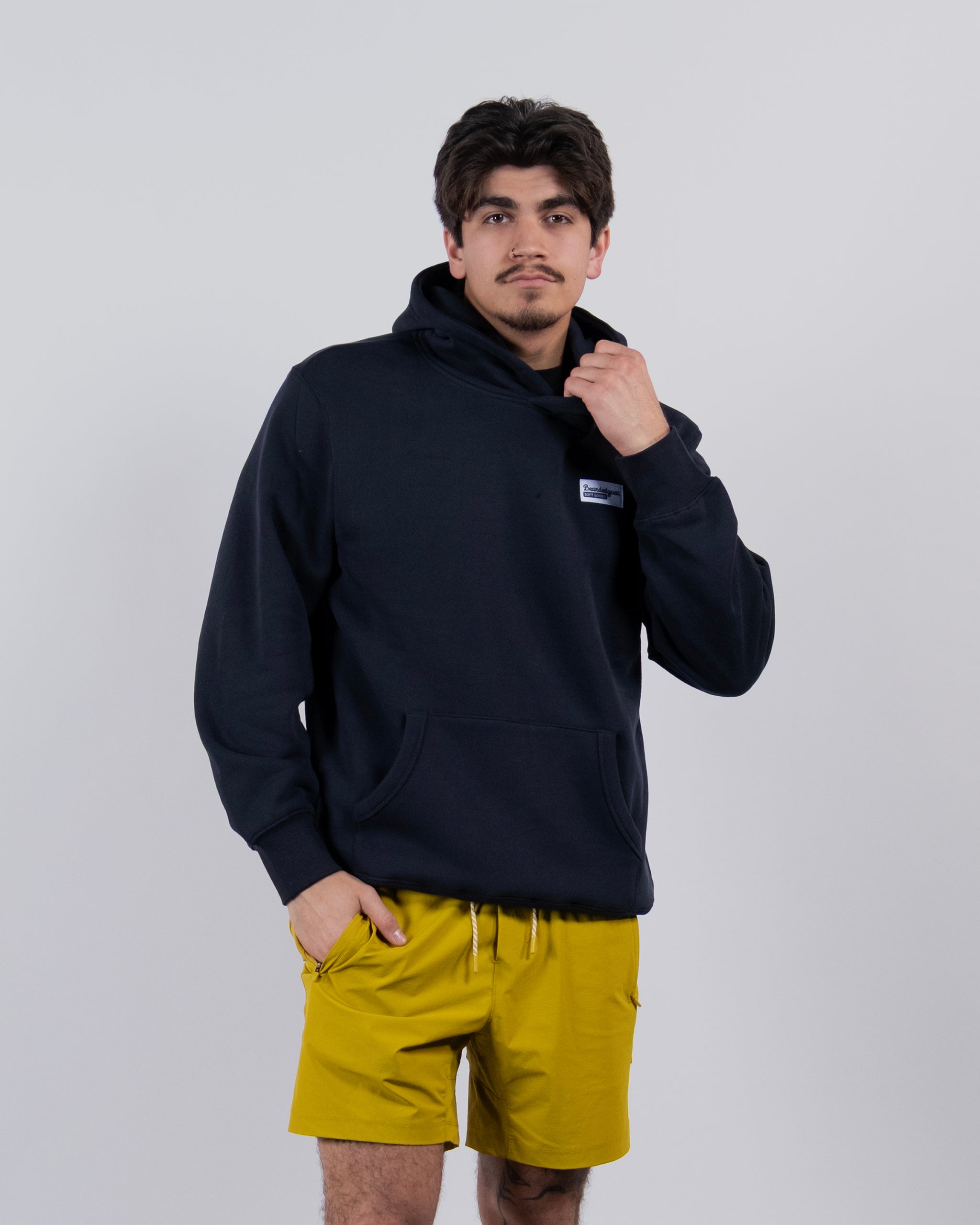 A man poses confidently against a plain background, wearing the best-selling beardedgoat The VOID™ Hoodie and bright yellow shorts, one hand in his pocket and the other adjusting his hoodie as he looks at the camera.