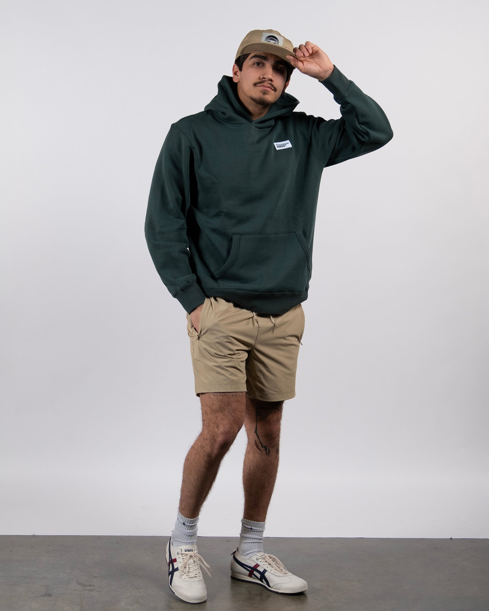 A man in a green beardedgoat The VOID™ Hoodie, khaki shorts, white socks and sneakers, and a beige cap stands against a plain background, touching the brim of his hat while looking at the camera.
