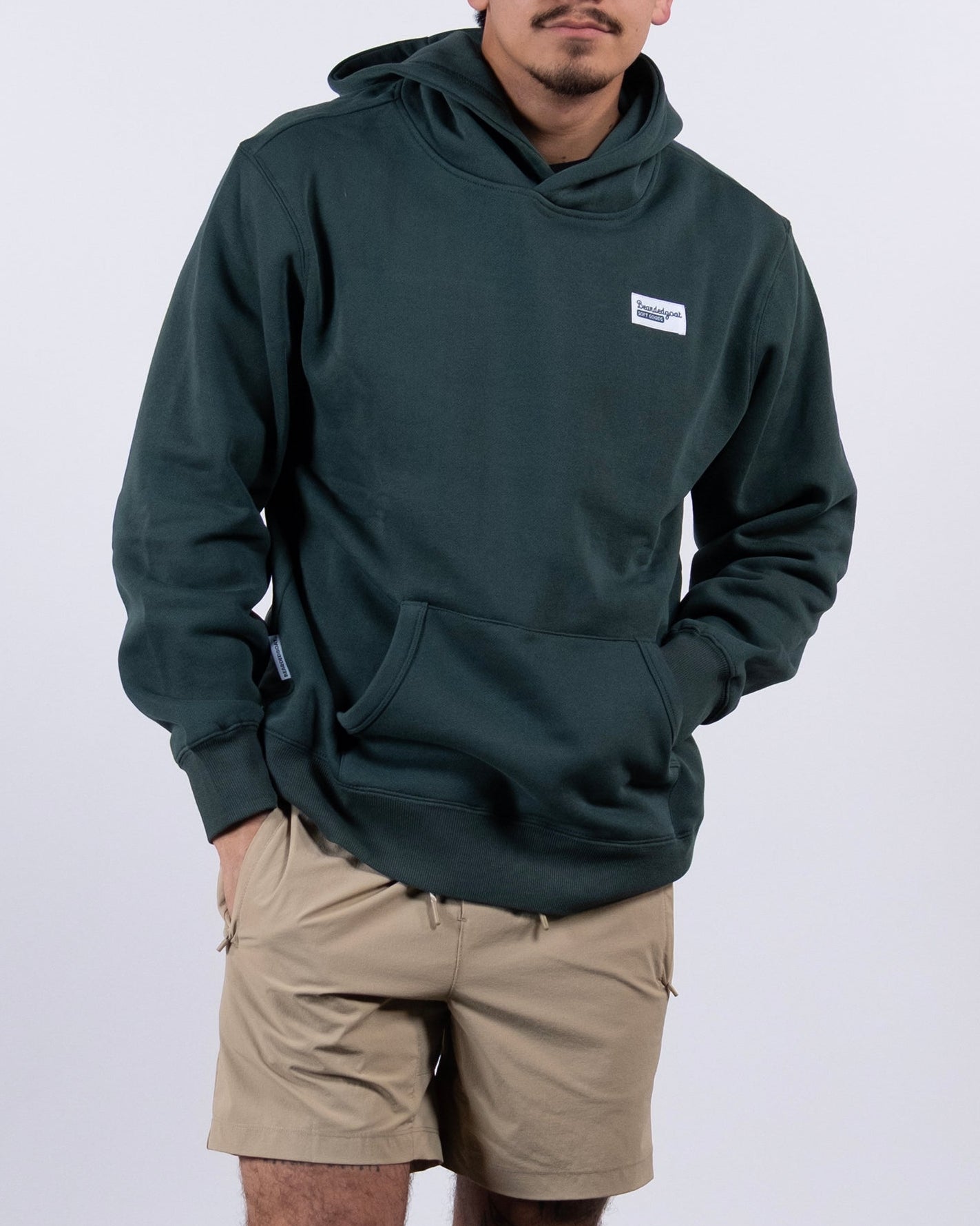 A man in a tan cap, tan shorts, and the beardedgoat The VOID™ Hoodie made from heavyweight cotton fleece stands against a plain white background with hands in his hoodie pocket, looking slightly to the side.