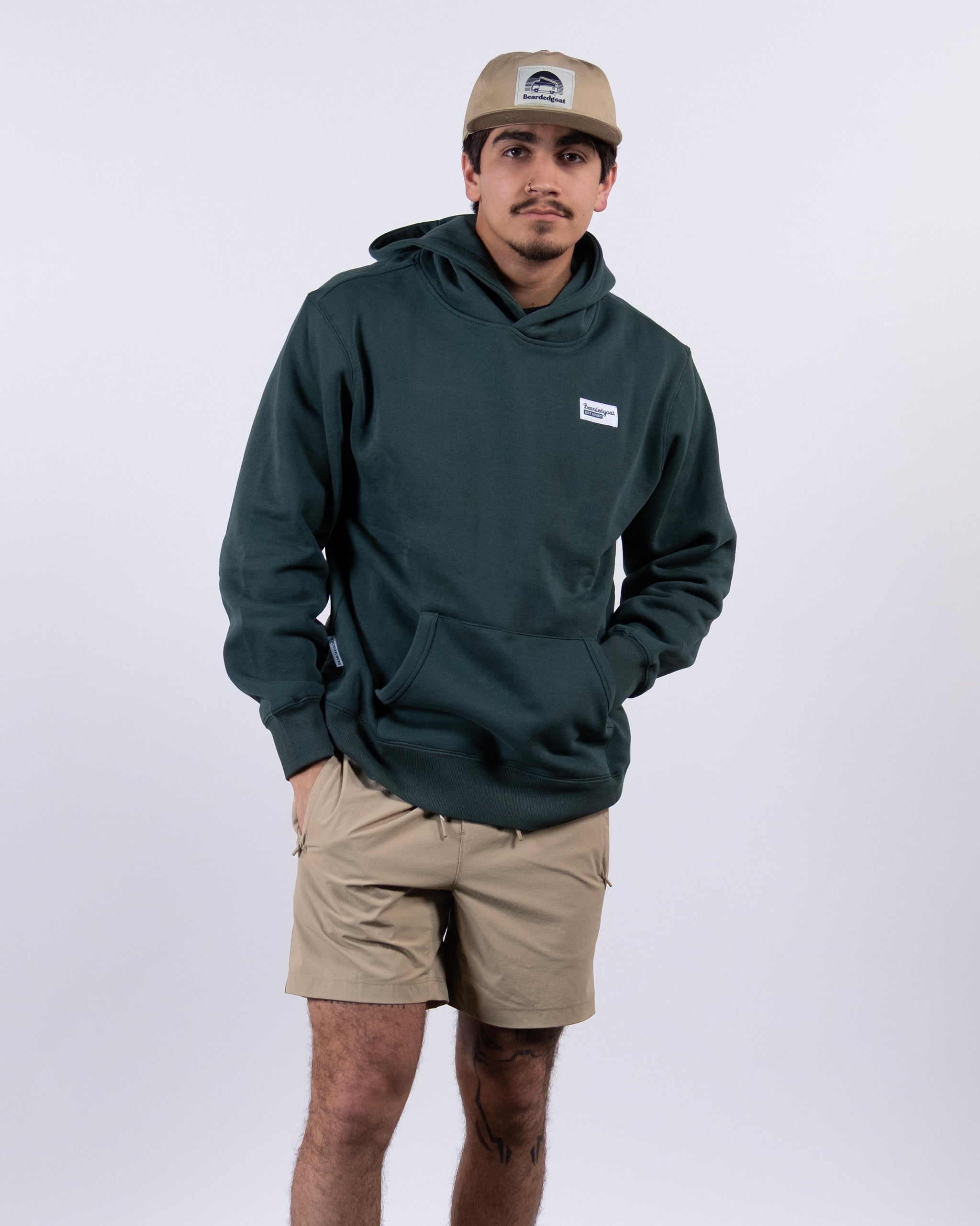 A man in a tan cap, tan shorts, and the beardedgoat The VOID™ Hoodie made from heavyweight cotton fleece stands against a plain white background with hands in his hoodie pocket, looking slightly to the side.