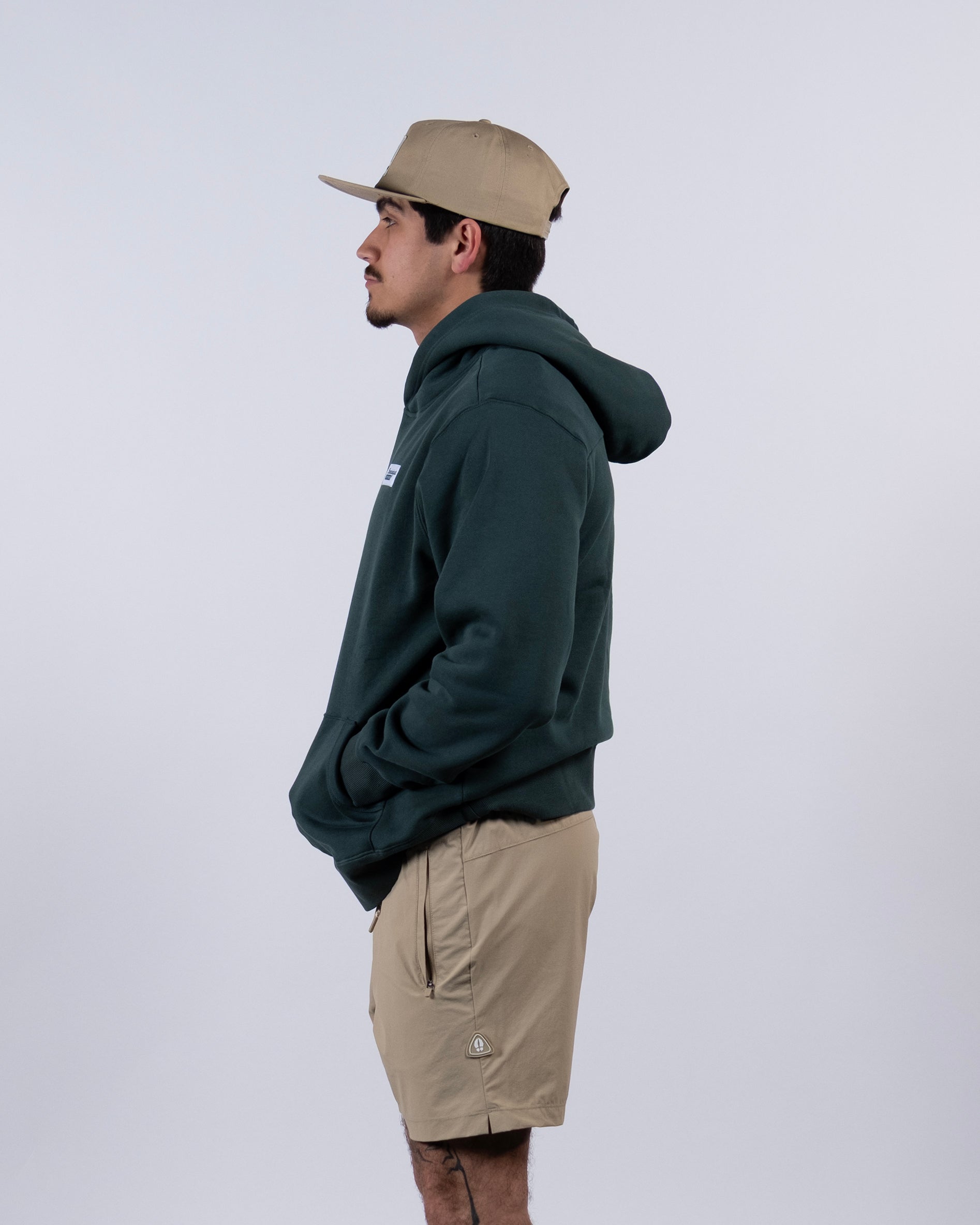 A man in profile wears tan shorts, a tan cap, and the beardedgoat VOID™ Hoodie—a dark green men's hoodie made from heavyweight cotton fleece—his hands tucked in the pockets, posing against a plain background.