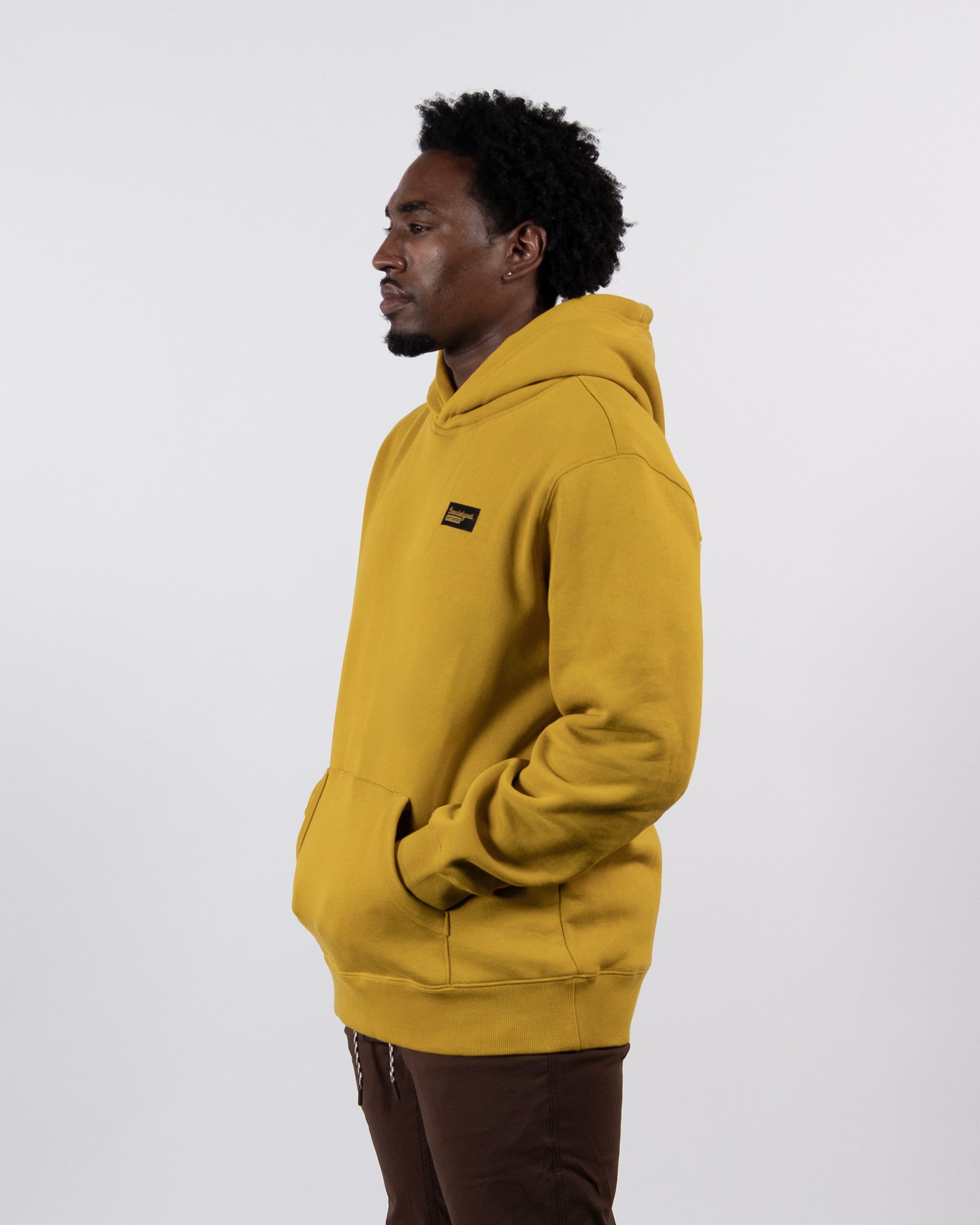 A person stands in profile against a plain background, wearing the beardedgoat The VOID™ Hoodie—crafted from premium heavyweight cotton fleece—with hands in the front pocket and brown pants.