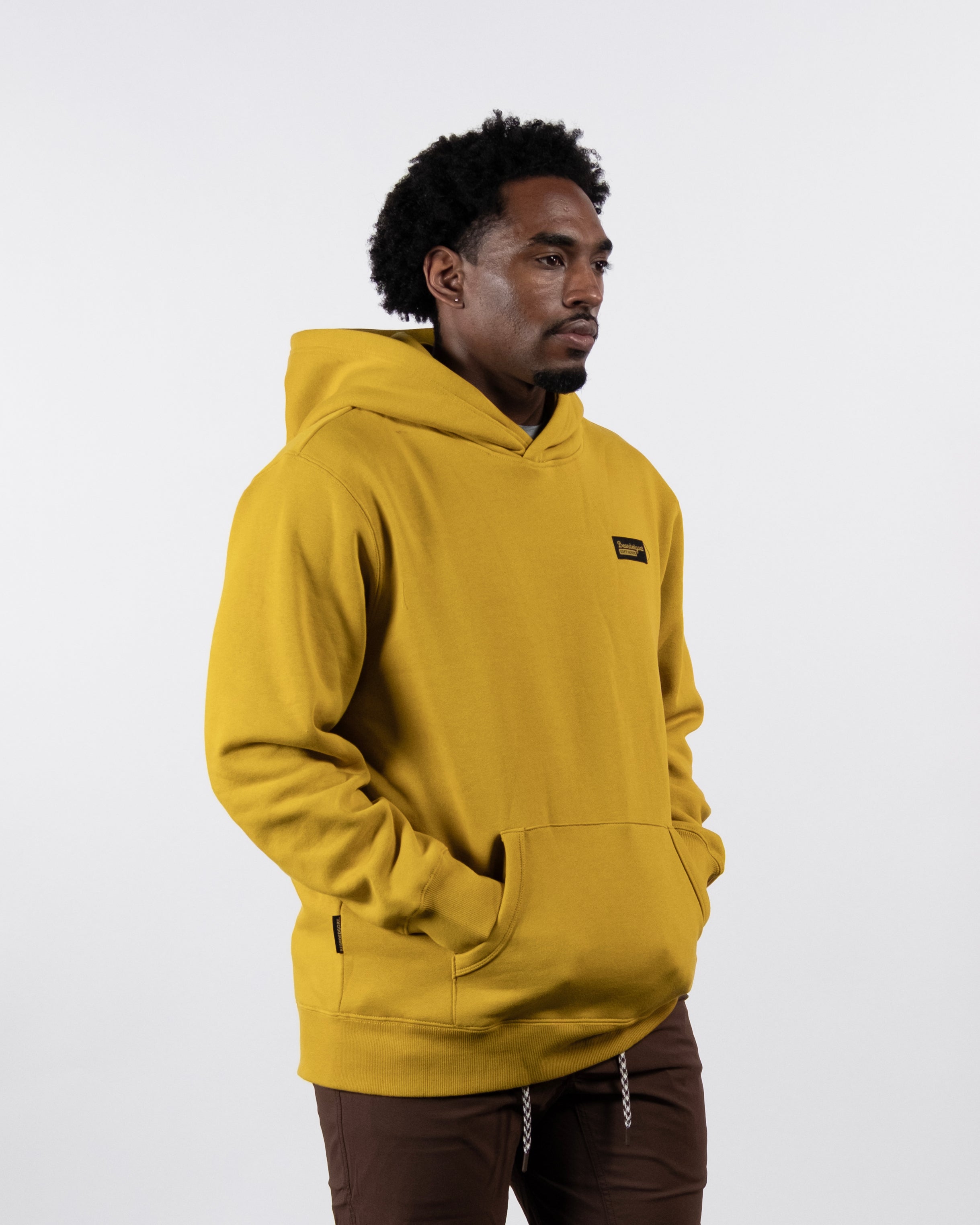 A man with short curly hair wears the beardedgoat VOID™ Hoodie in mustard yellow and brown pants made from heavyweight cotton fleece, standing against a plain white background with his hands in the hoodie’s front pocket.