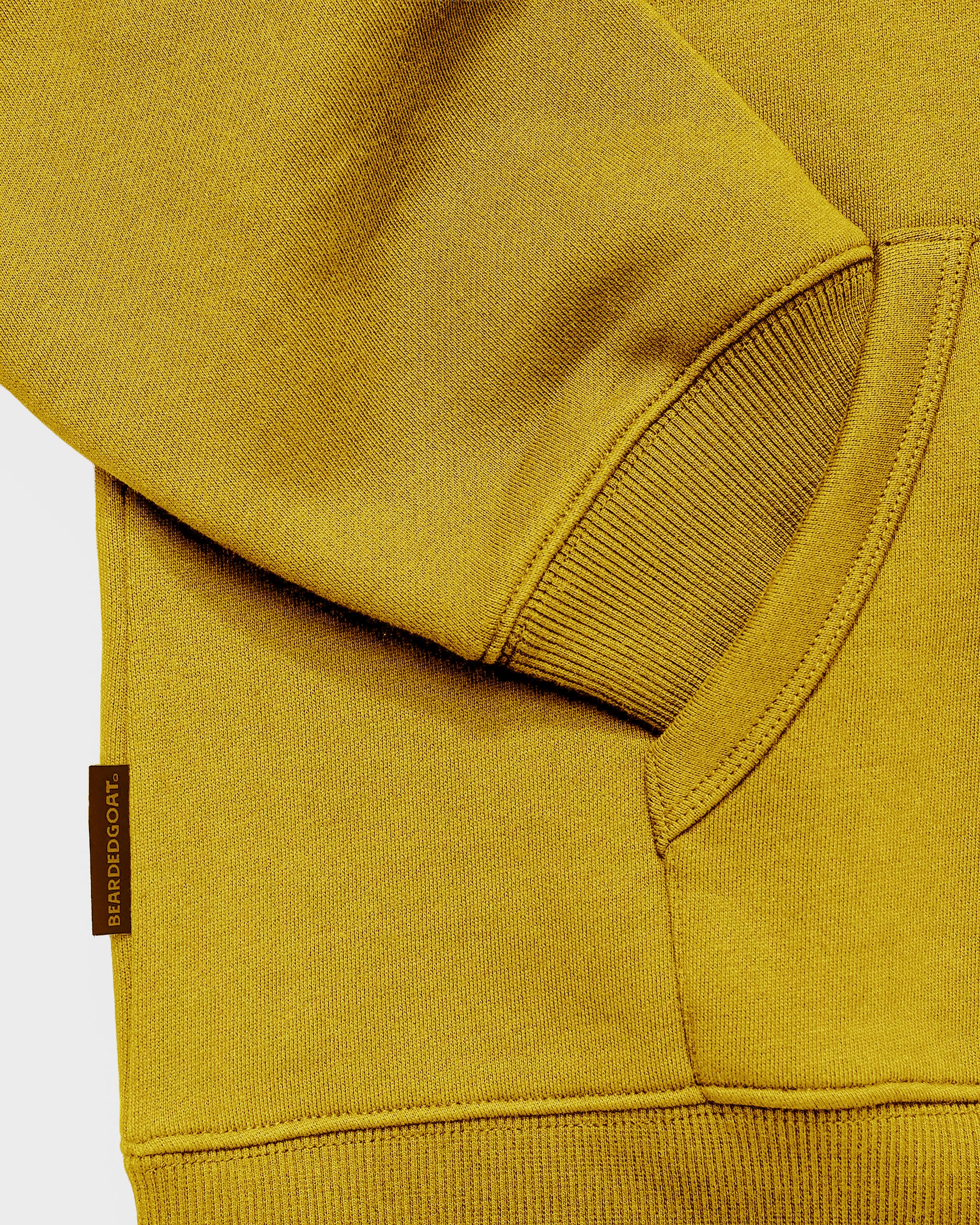 A close-up of the beardedgoat The VOID™ Hoodie in mustard yellow highlights its ribbed cuff, pocket, brown “HARDGRAFT” tag, and soft, heavyweight cotton fleece fabric.