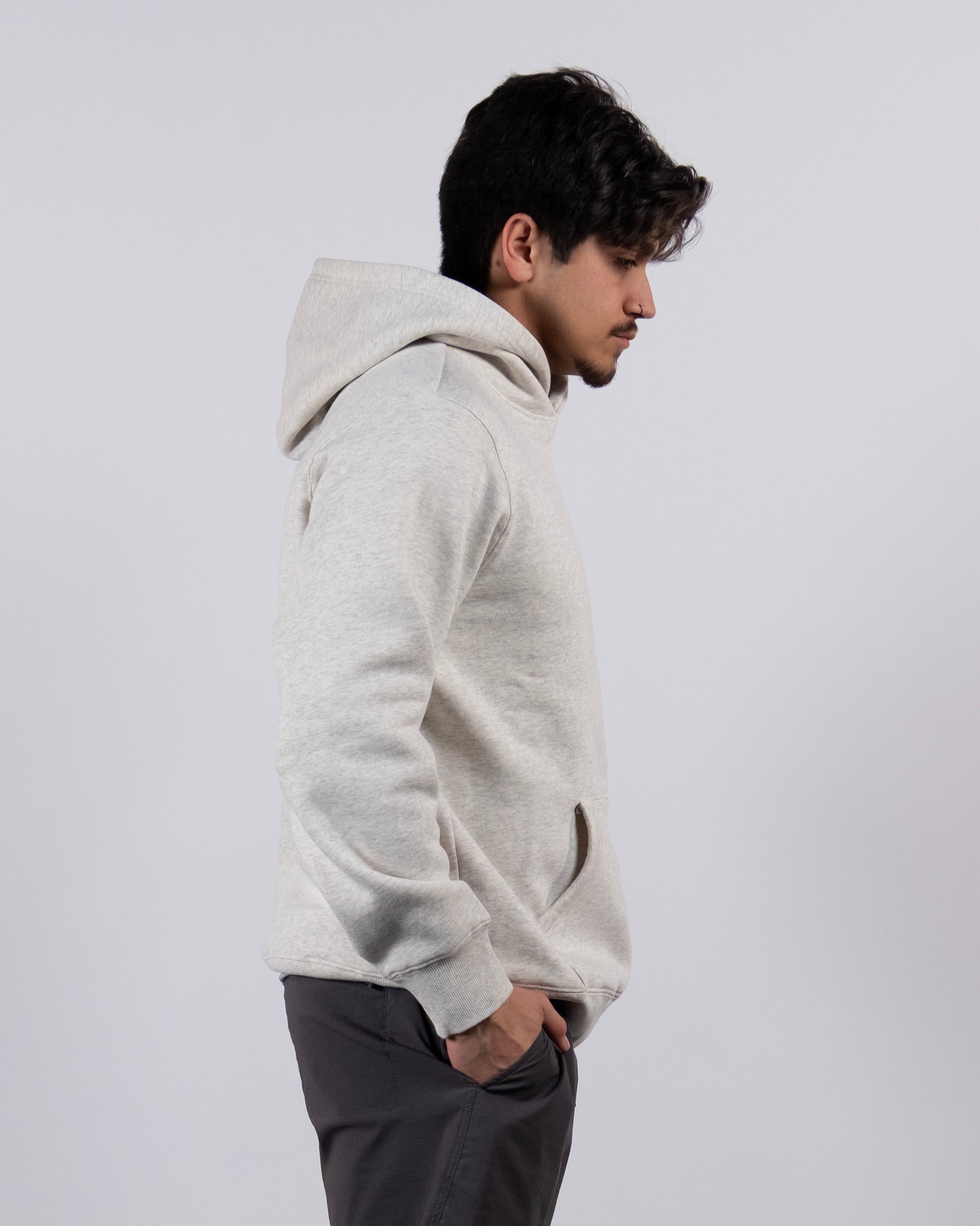 A man wears The VOID™ Hoodie by beardedgoat—a light gray, heavyweight cotton fleece men's hoodie—with dark pants, standing in profile against a plain light background, hands in his pockets and looking slightly downward.