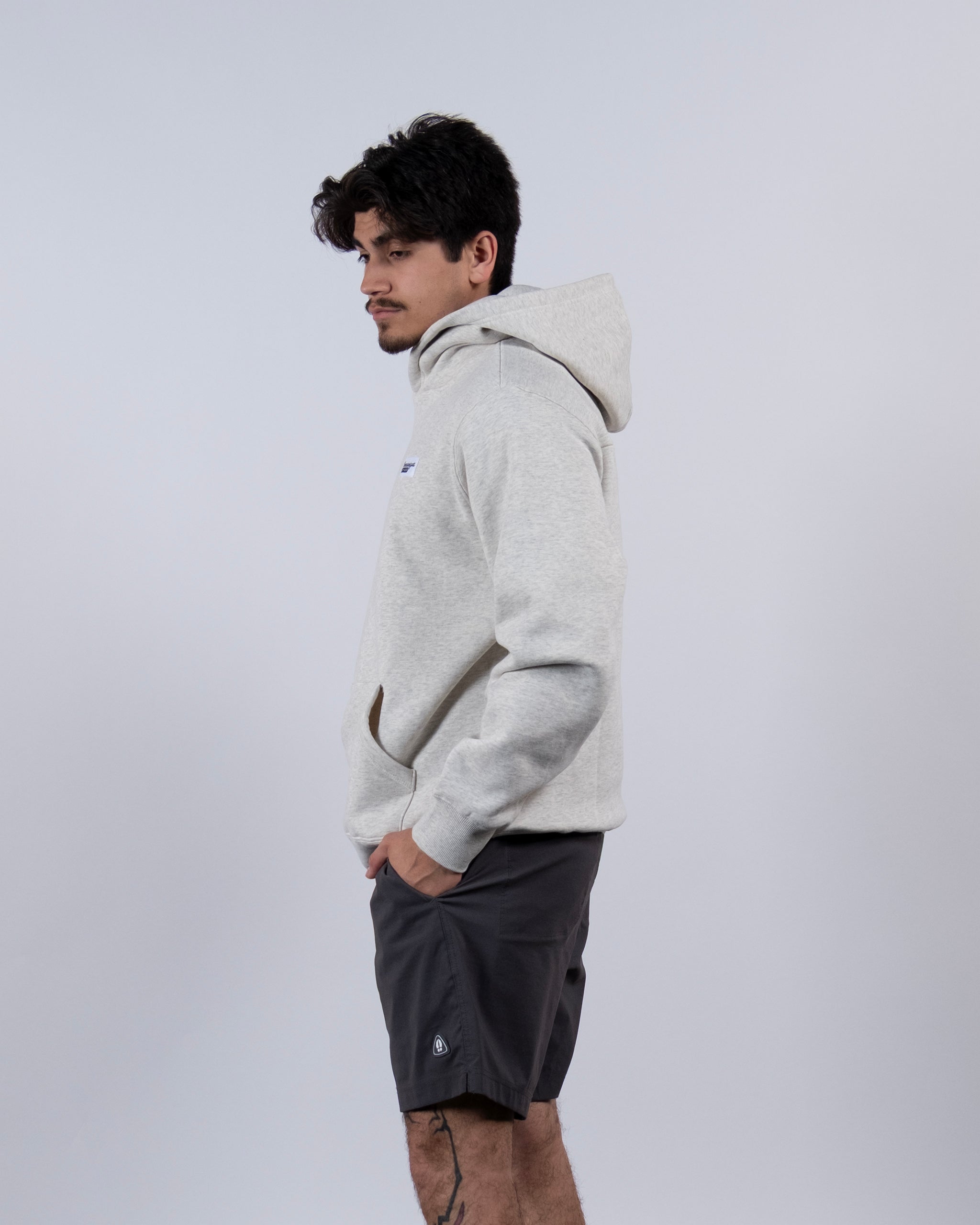 A person stands sideways with hands in pockets, wearing a light gray beardedgoat The VOID™ Hoodie made from heavyweight cotton fleece and dark shorts, set against a plain light gray background.