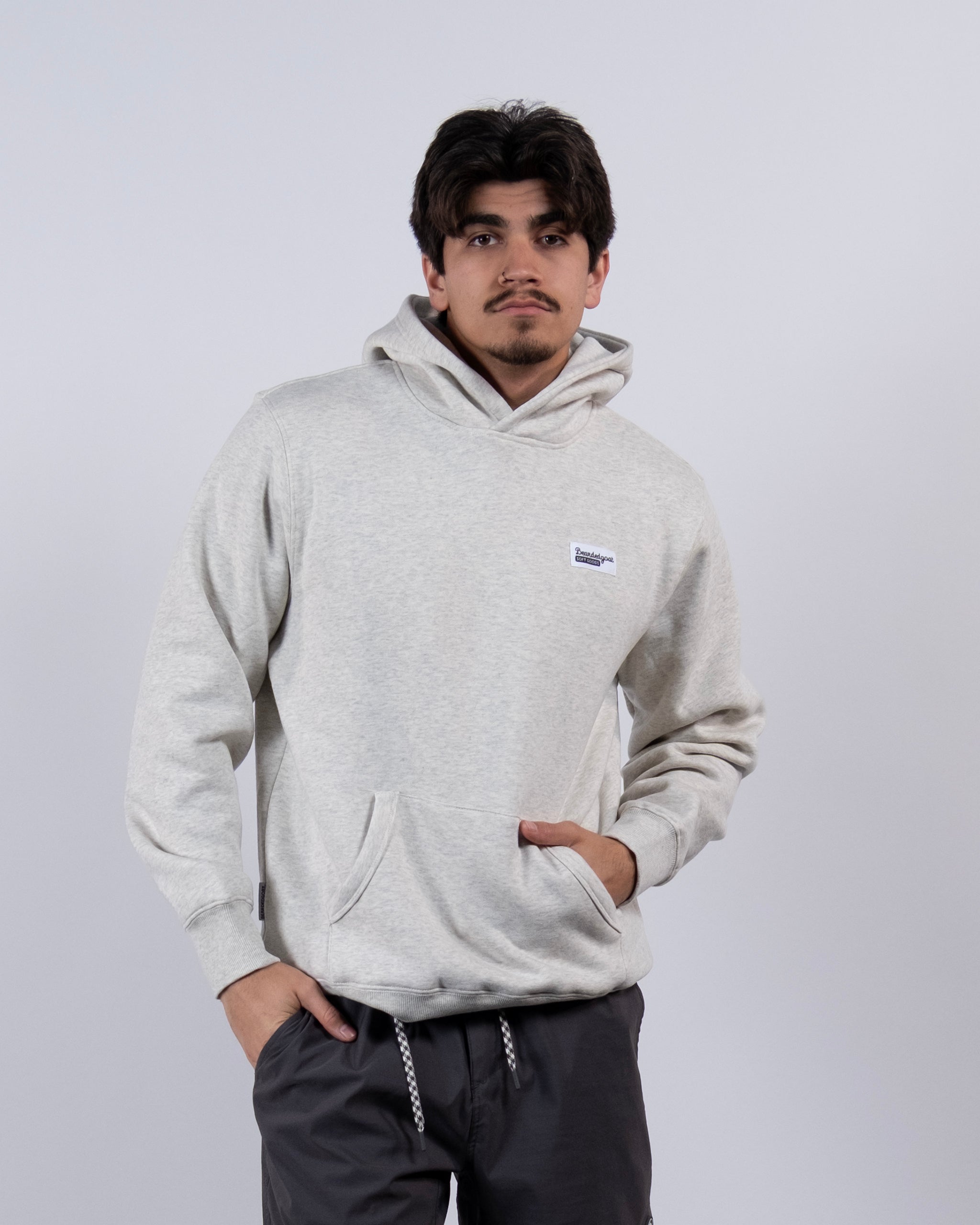 A young man with short dark hair wears The VOID™ Hoodie by beardedgoat, made of heavyweight cotton fleece, and dark gray drawstring pants. He stands against a plain light background with one hand in his hoodie pocket and a neutral expression.