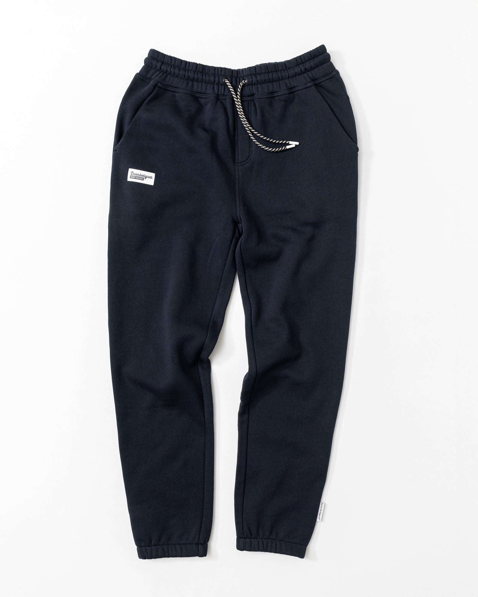 Men's Void Sweatpants by beardedgoat in navy blue, featuring an elastic waistband with drawstring, side pockets, a zippered back pocket, a white patch on the left thigh, and elastic ankle cuffs, shown laid flat on a white background.
