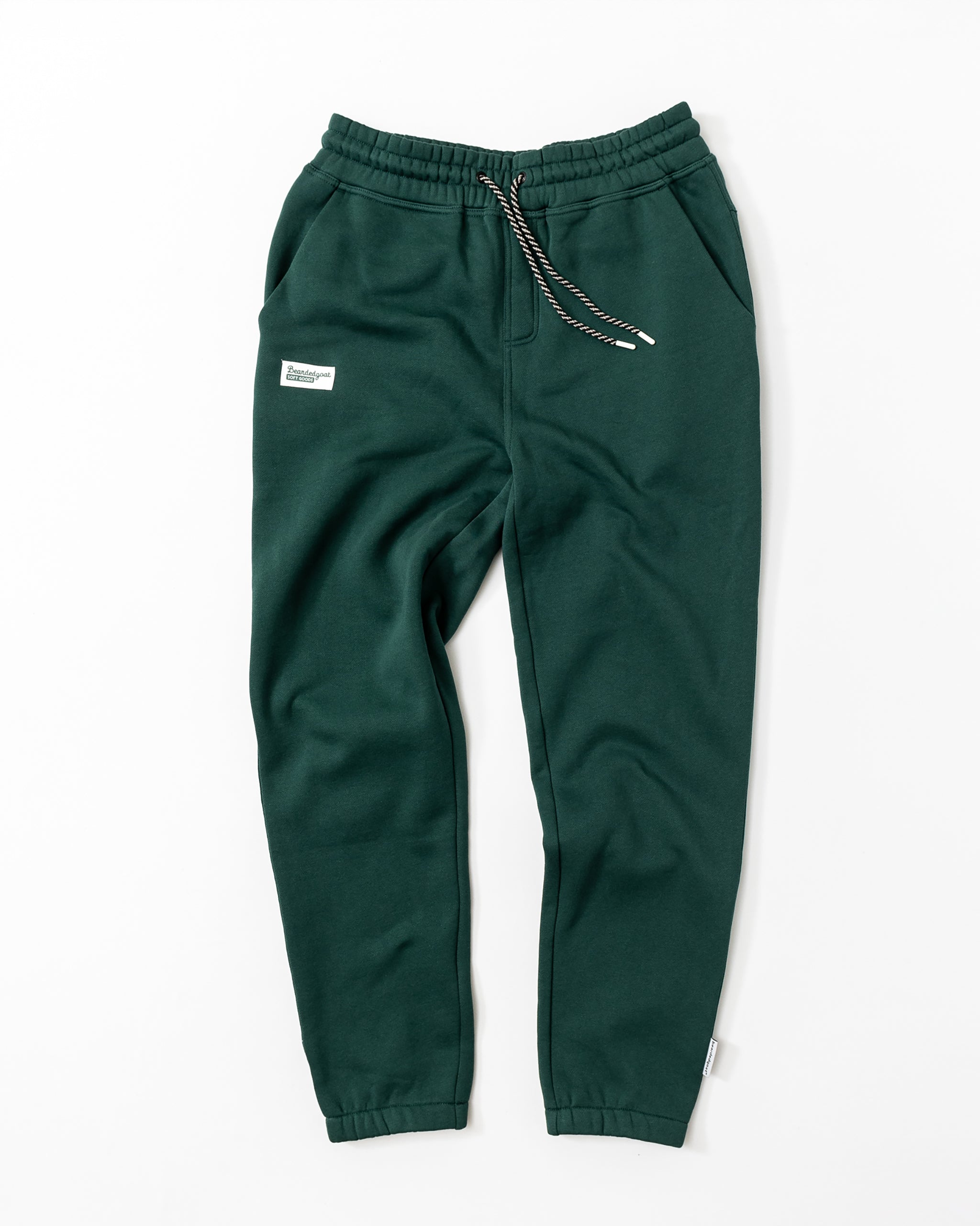 Men's Void Sweatpants by beardedgoat in dark green, featuring an elastic waistband, black-and-white drawstring, side pockets, a zippered back pocket, and a small white label on the left hip. Laid flat on a white background.