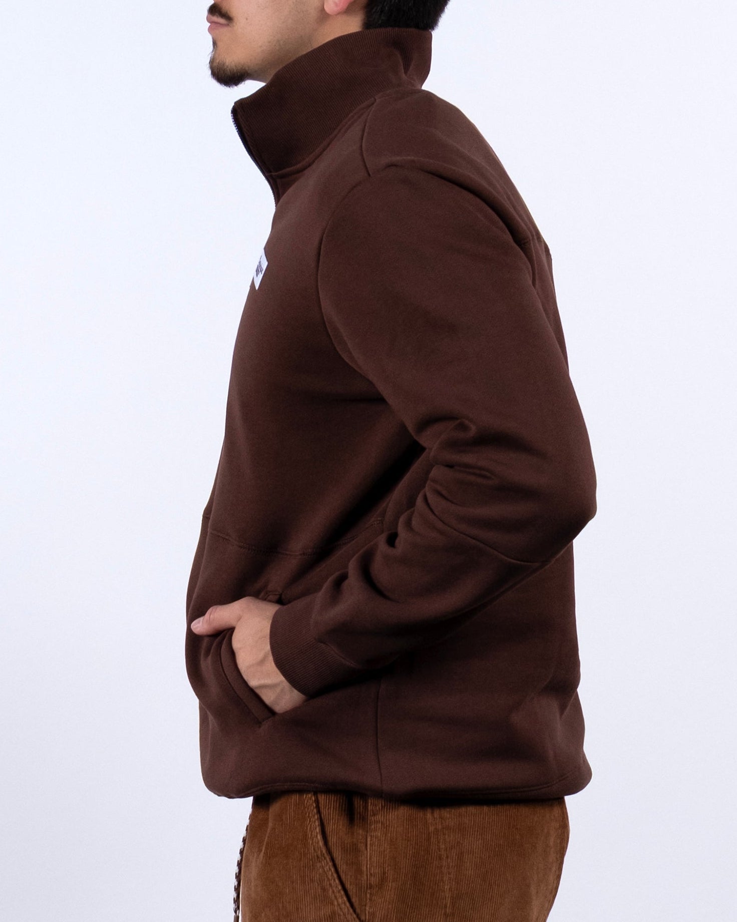 A man with dark hair and a mustache stands in profile against a white background, wearing the BEARDEDGOAT NEW — VOID™ Half Zip jacket with matching pants, his hands tucked into the jacket pockets.