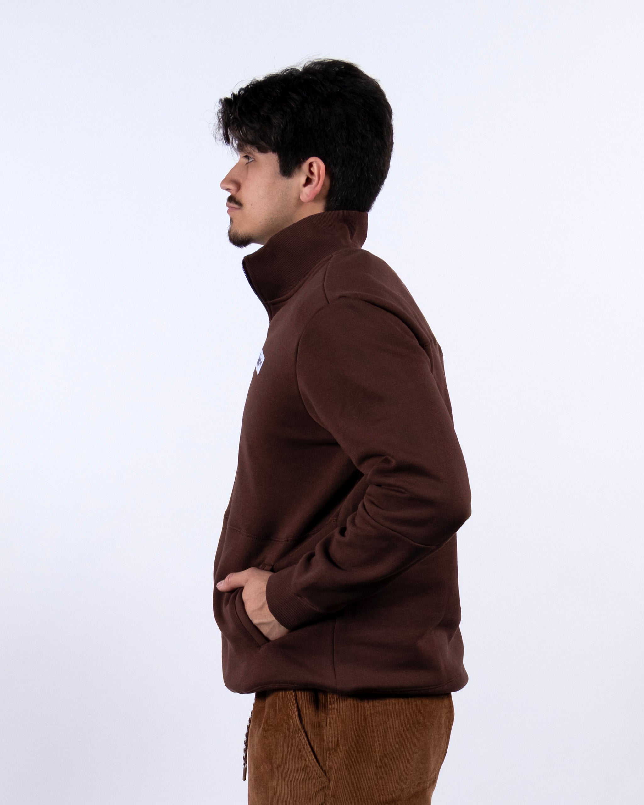 A man with dark hair and a mustache stands in profile against a white background, wearing the BEARDEDGOAT NEW — VOID™ Half Zip jacket with matching pants, his hands tucked into the jacket pockets.