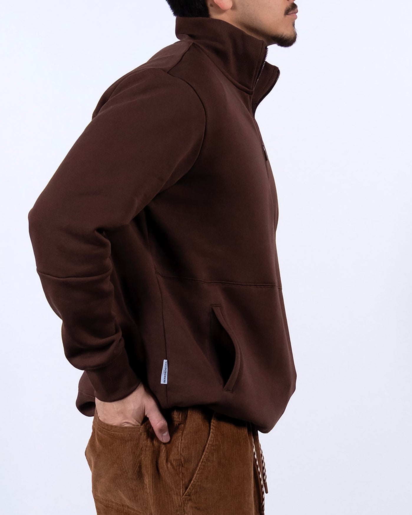 A man with short dark hair, wearing the BEARDEDGOAT NEW — VOID™ Half Zip brown jacket and brown corduroy pants, stands in profile against a plain white background with his hands in his pockets.