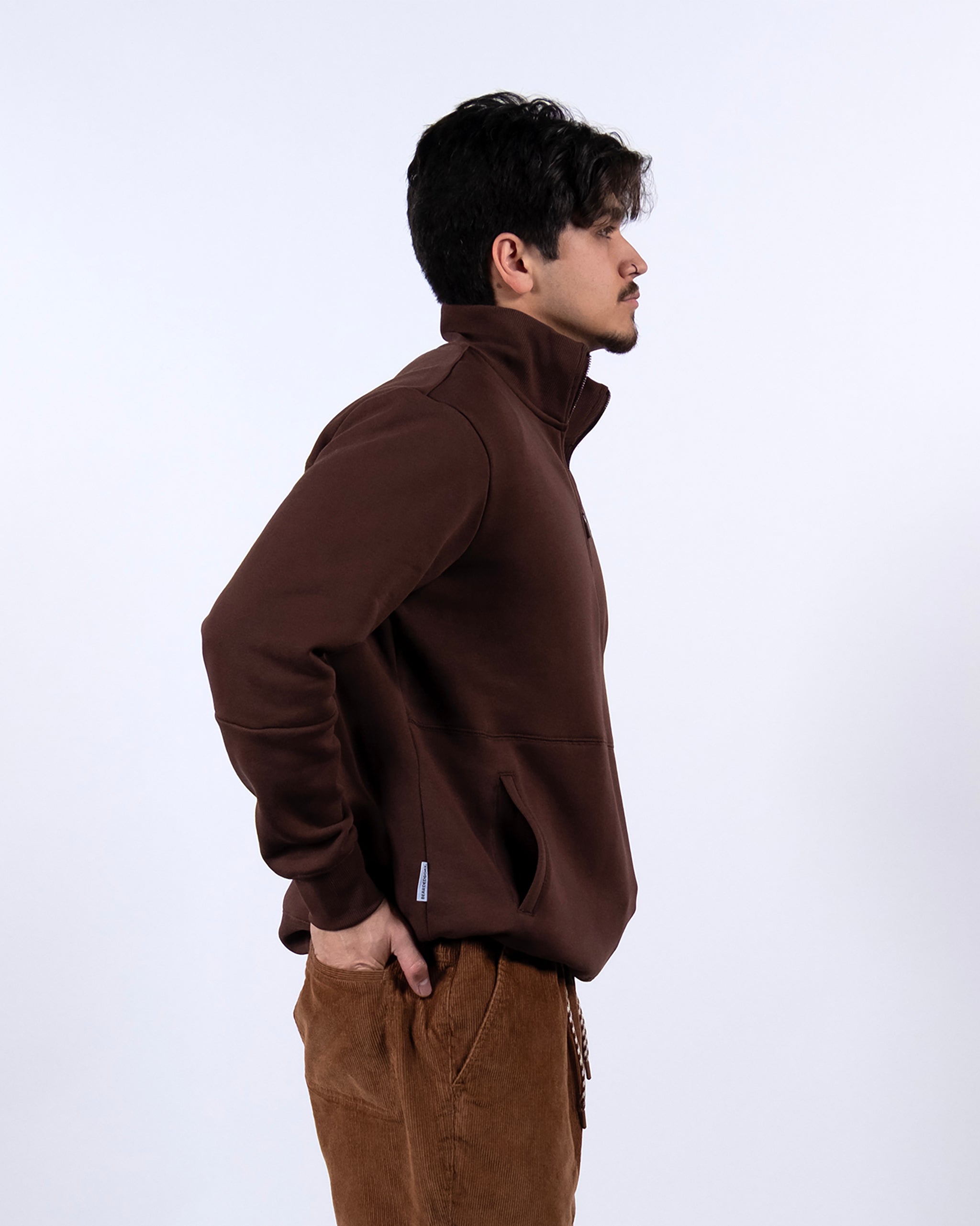 A man with short dark hair, wearing the BEARDEDGOAT NEW — VOID™ Half Zip brown jacket and brown corduroy pants, stands in profile against a plain white background with his hands in his pockets.
