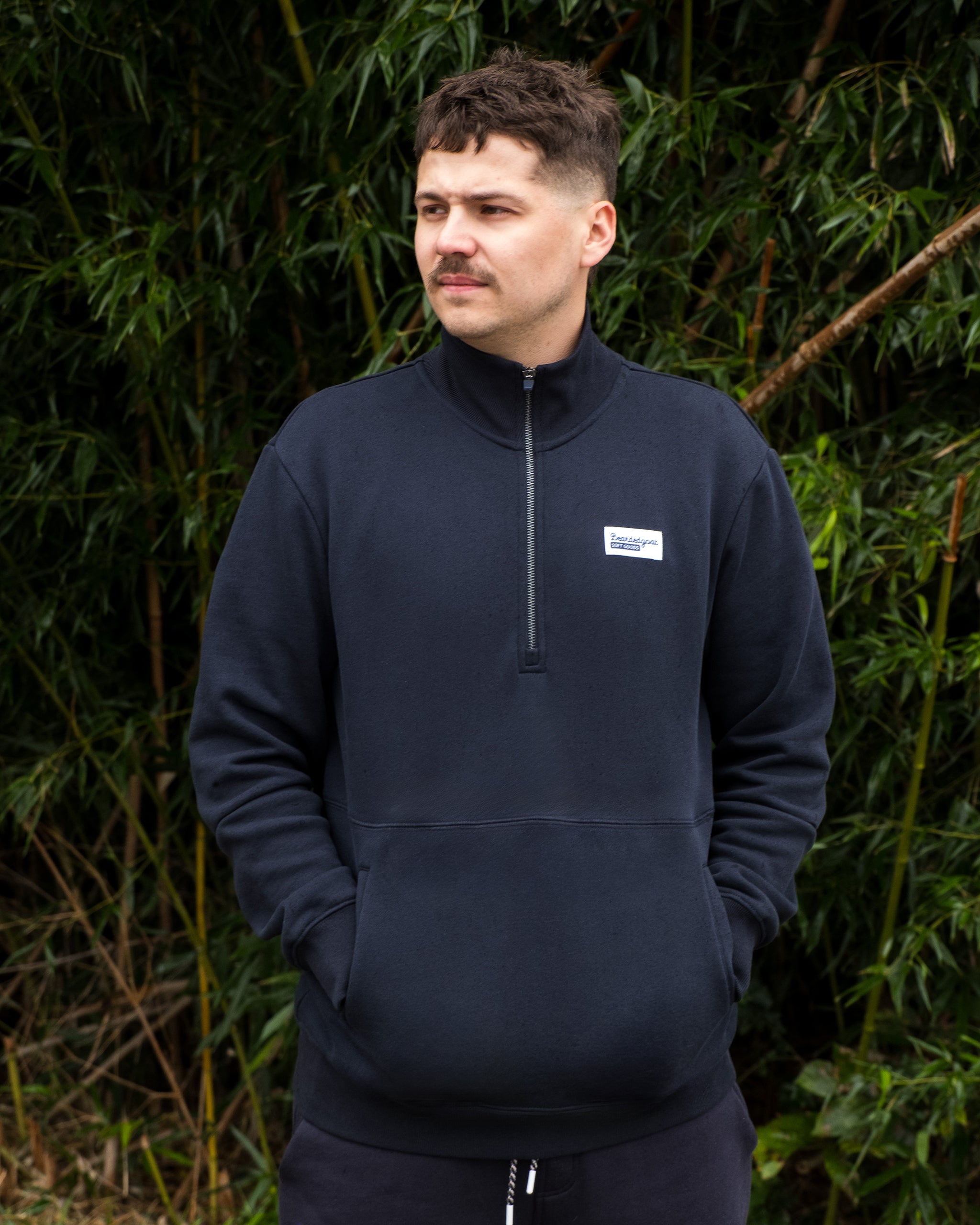 A person with short dark hair and a mustache stands outdoors before green foliage, wearing the BEARDEDGOAT NEW — VOID™ Half Zip with hands in the front pocket, looking to the side.