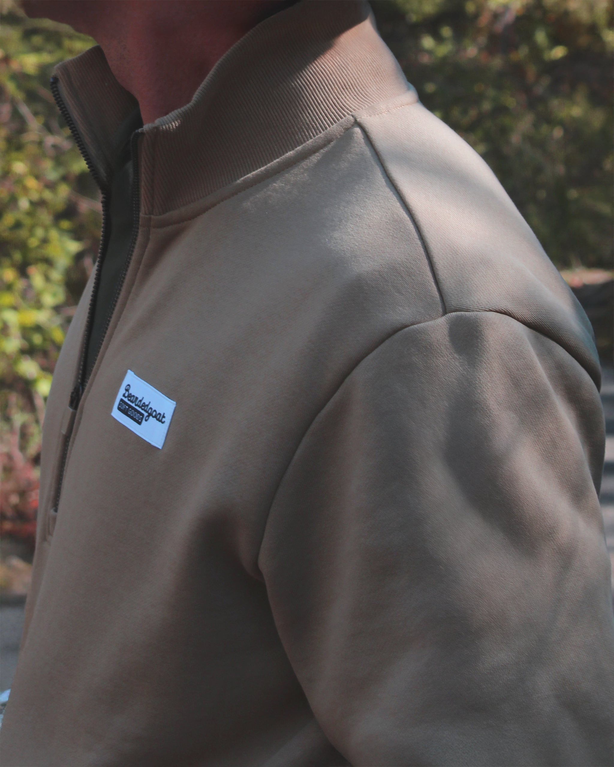 A person wearing the BEARDEDGOAT NEW — VOID™ Half Zip premium brown cotton fleece jacket with a small rectangular white and blue chest patch stands outdoors, greenery in the background.