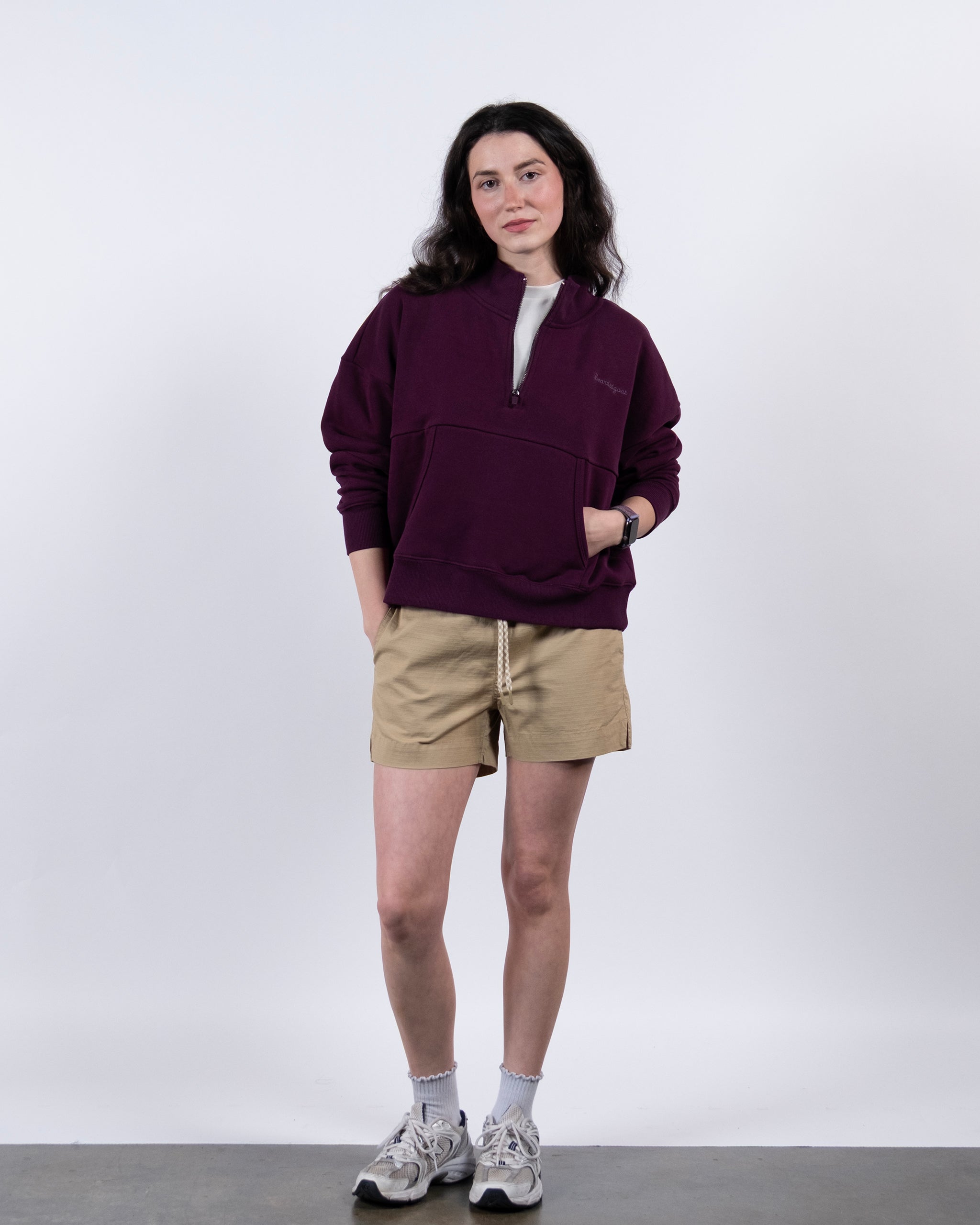 A woman stands against a white background wearing the BEARDEDGOAT NEW — Overnight™ Half Zip in maroon, paired with beige shorts, white socks, and gray sneakers. Her hands are in her pockets as she looks at the camera with a neutral expression.