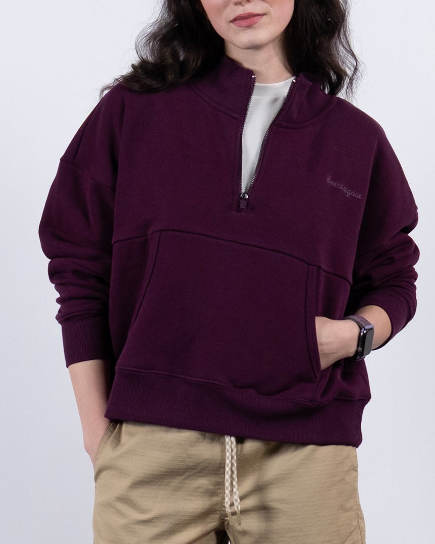 A woman stands against a white background wearing the BEARDEDGOAT NEW — Overnight™ Half Zip in maroon, paired with beige shorts, white socks, and gray sneakers. Her hands are in her pockets as she looks at the camera with a neutral expression.