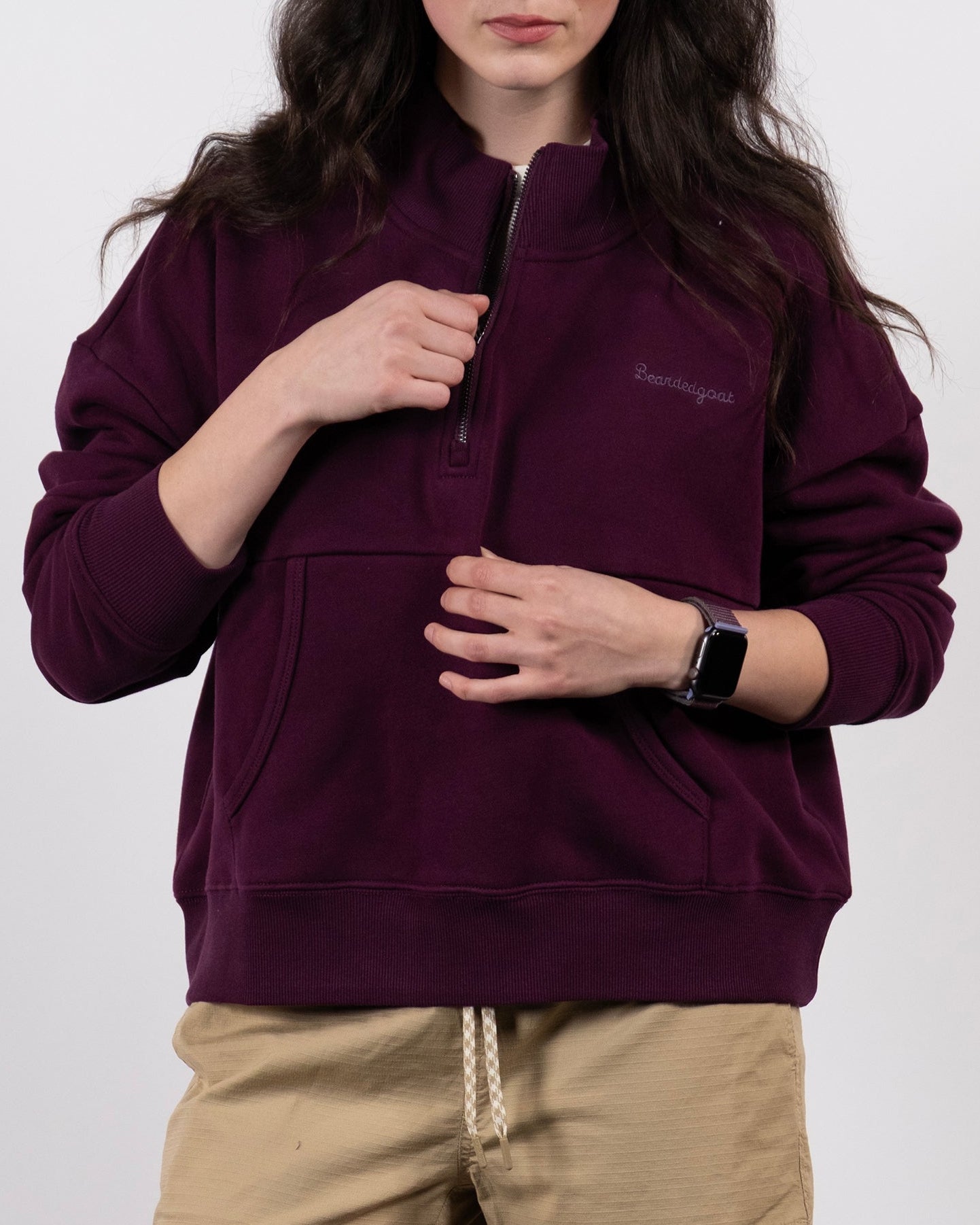 A person with long dark hair wears the BEARDEDGOAT NEW — Overnight™ Half Zip in maroon and khaki shorts, standing against a plain background while looking down and holding the zipper.