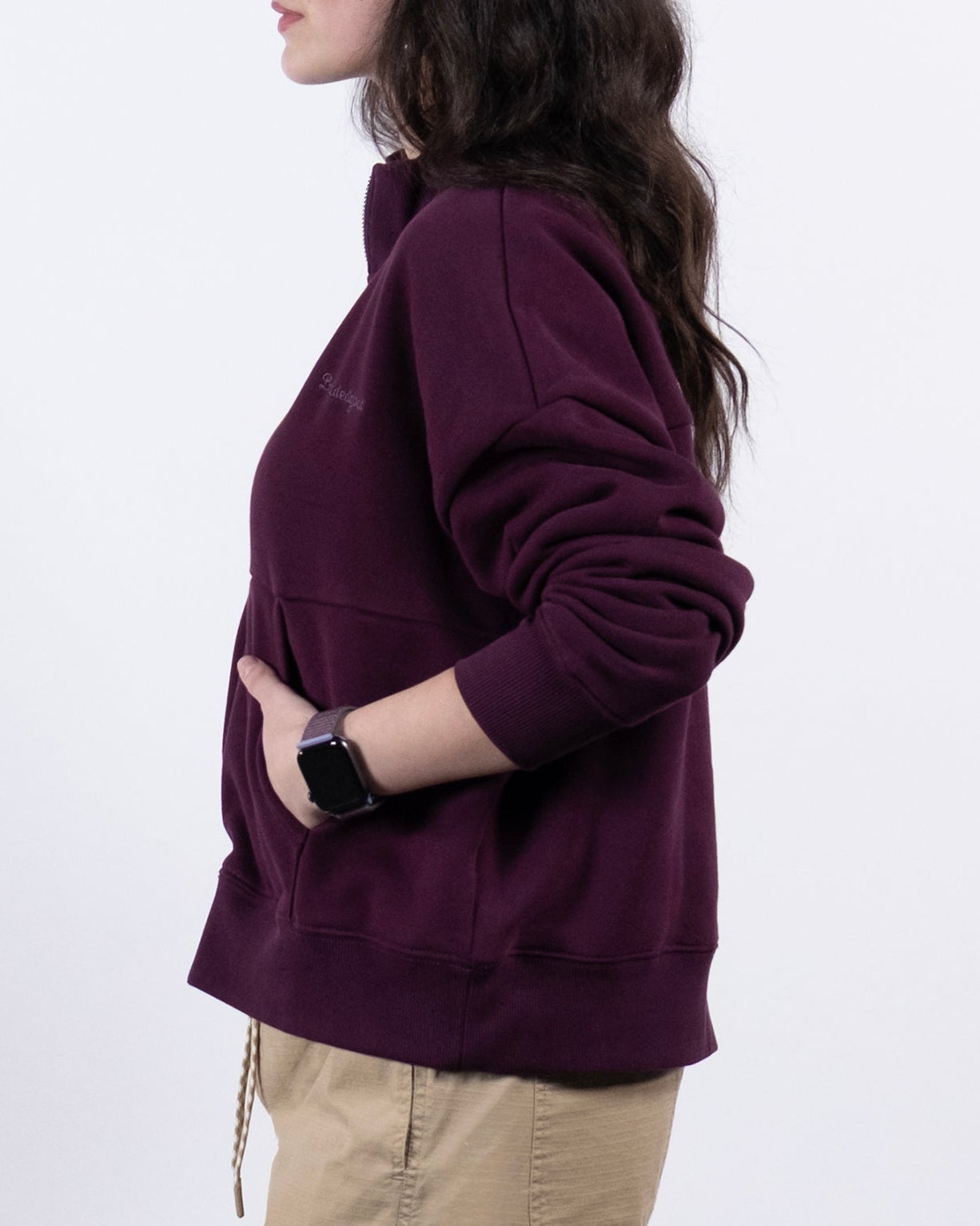 A woman with long dark hair stands in profile against a white background, wearing the BEARDEDGOAT NEW — Overnight™ Half Zip maroon heavyweight fleece sweatshirt, beige shorts, and a smartwatch. Her hands are in the sweatshirt pocket.