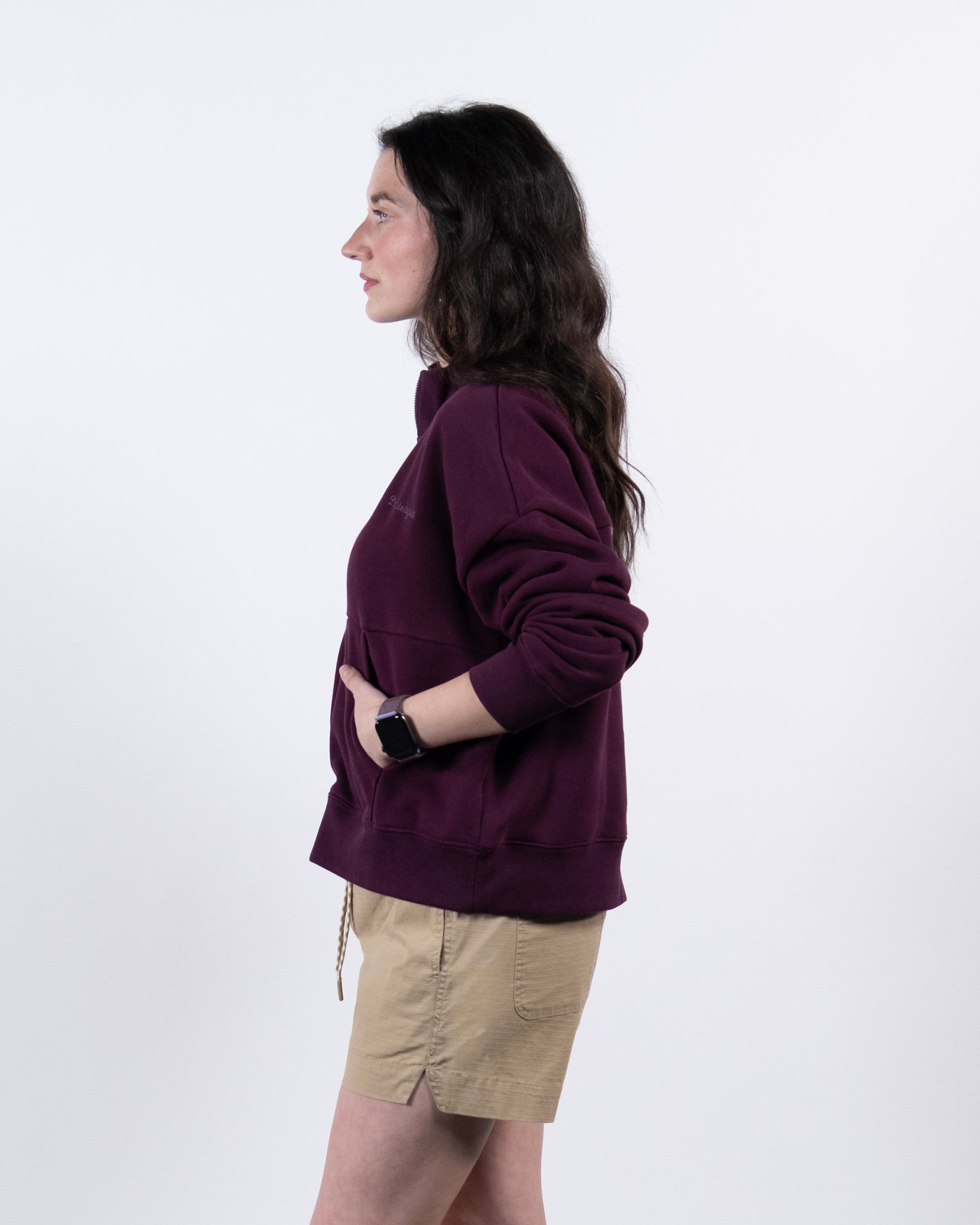 A woman with long dark hair stands in profile against a white background, wearing the BEARDEDGOAT NEW — Overnight™ Half Zip maroon heavyweight fleece sweatshirt, beige shorts, and a smartwatch. Her hands are in the sweatshirt pocket.