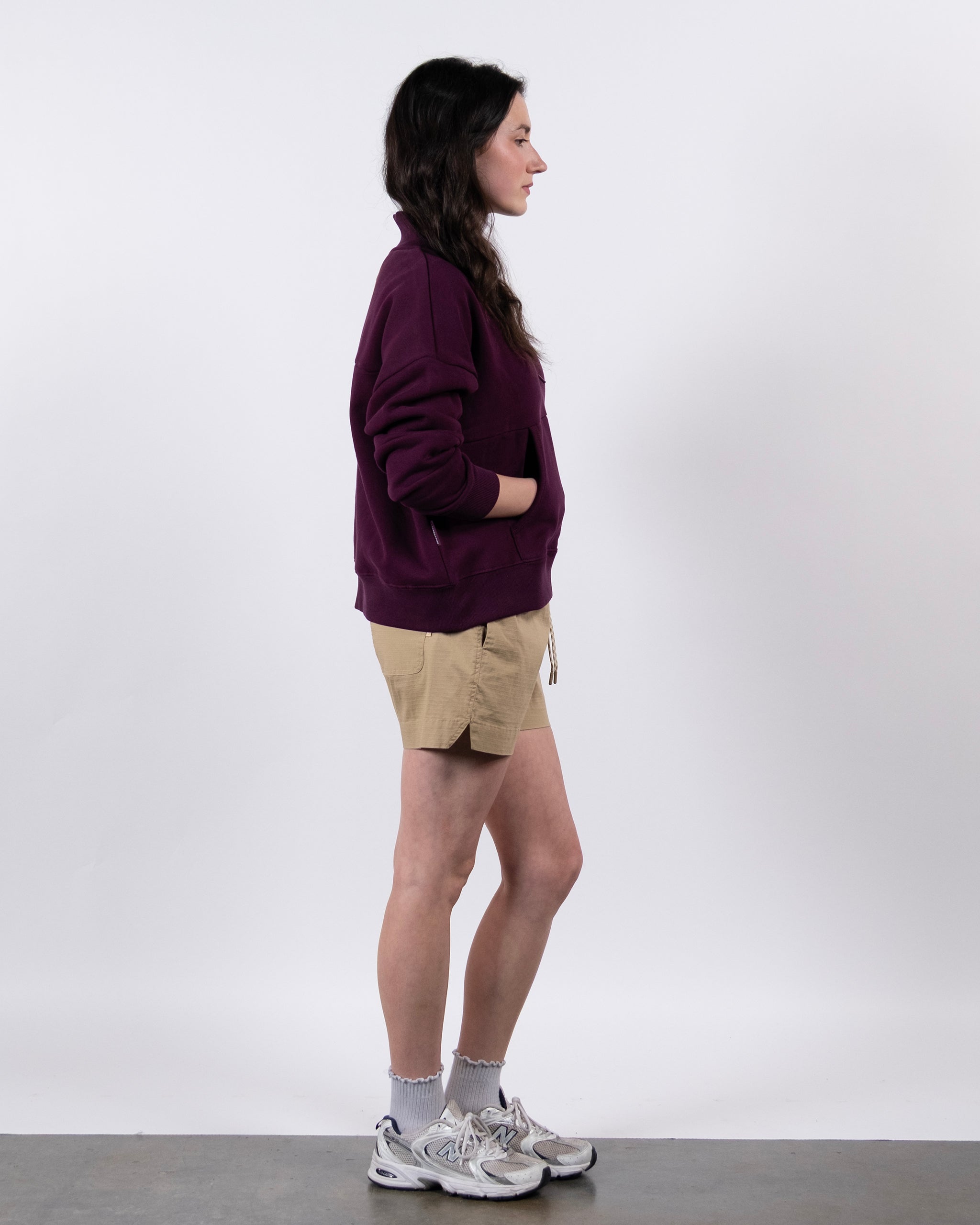 A woman stands in profile against a white background, wearing the BEARDEDGOAT NEW — Overnight™ Half Zip in maroon, tan shorts, gray socks, and white sneakers, with her hands in the sweatshirt's pockets.