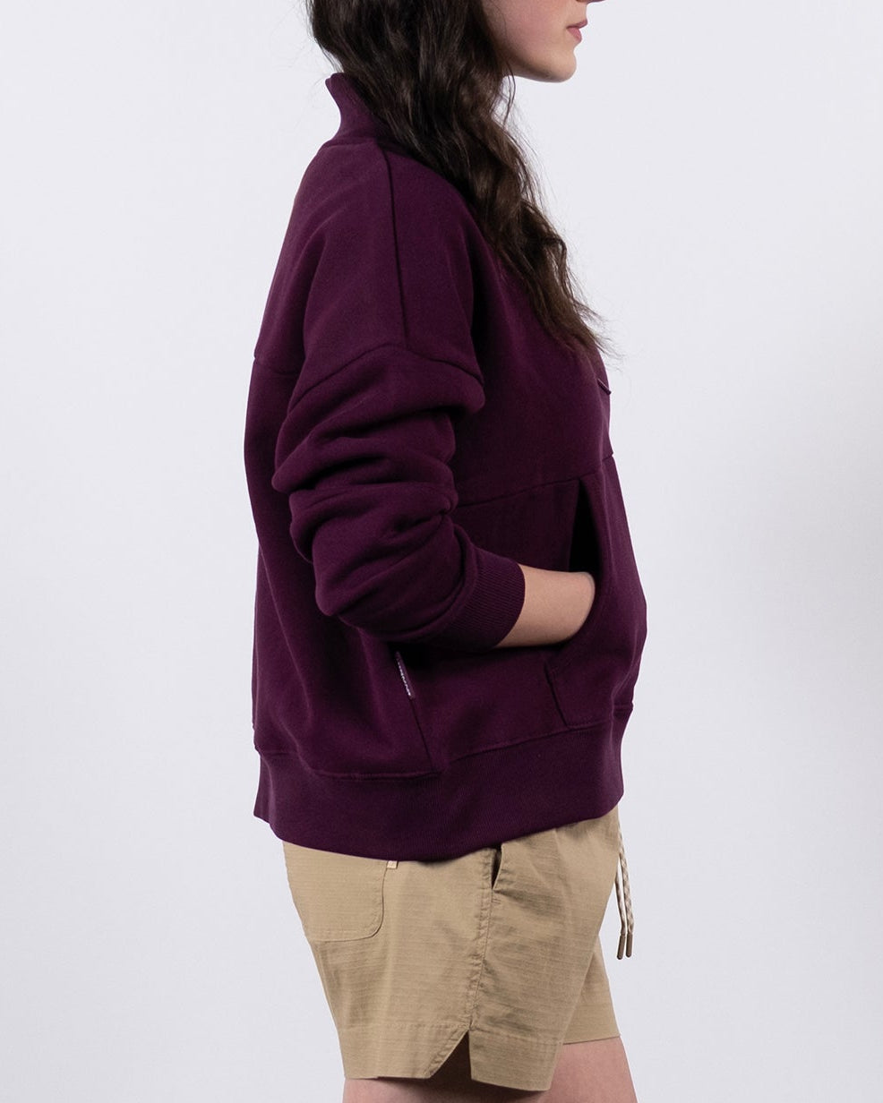 A woman stands in profile against a white background, wearing the BEARDEDGOAT NEW — Overnight™ Half Zip in maroon, tan shorts, gray socks, and white sneakers, with her hands in the sweatshirt's pockets.