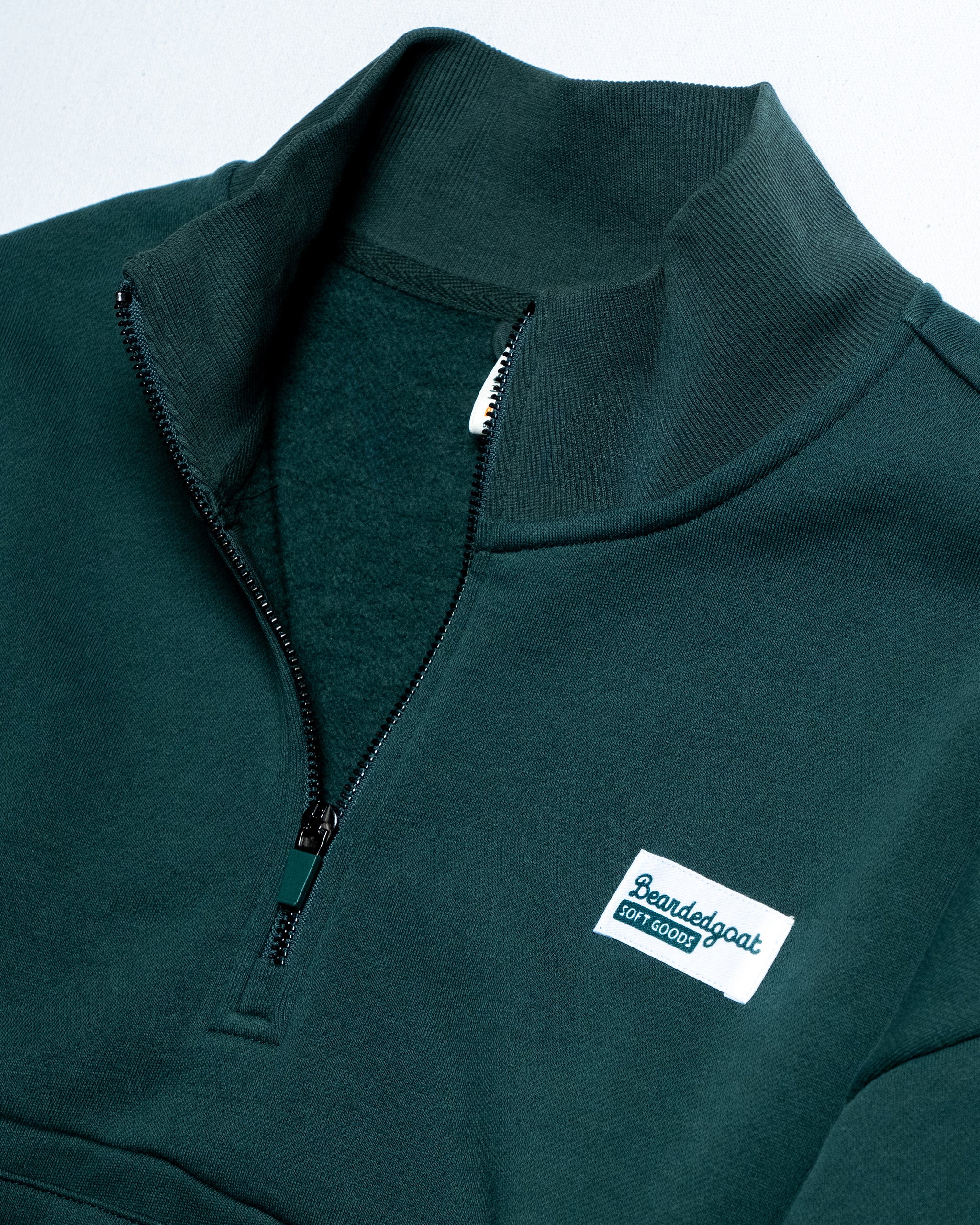 The BEARDEDGOAT NEW — Overnight™ Half Zip is a dark green cotton fleece quarter-zip sweater with a ribbed collar, partially unzipped, and a white rectangular Boardegoat Sport Goods logo patch on the left chest.