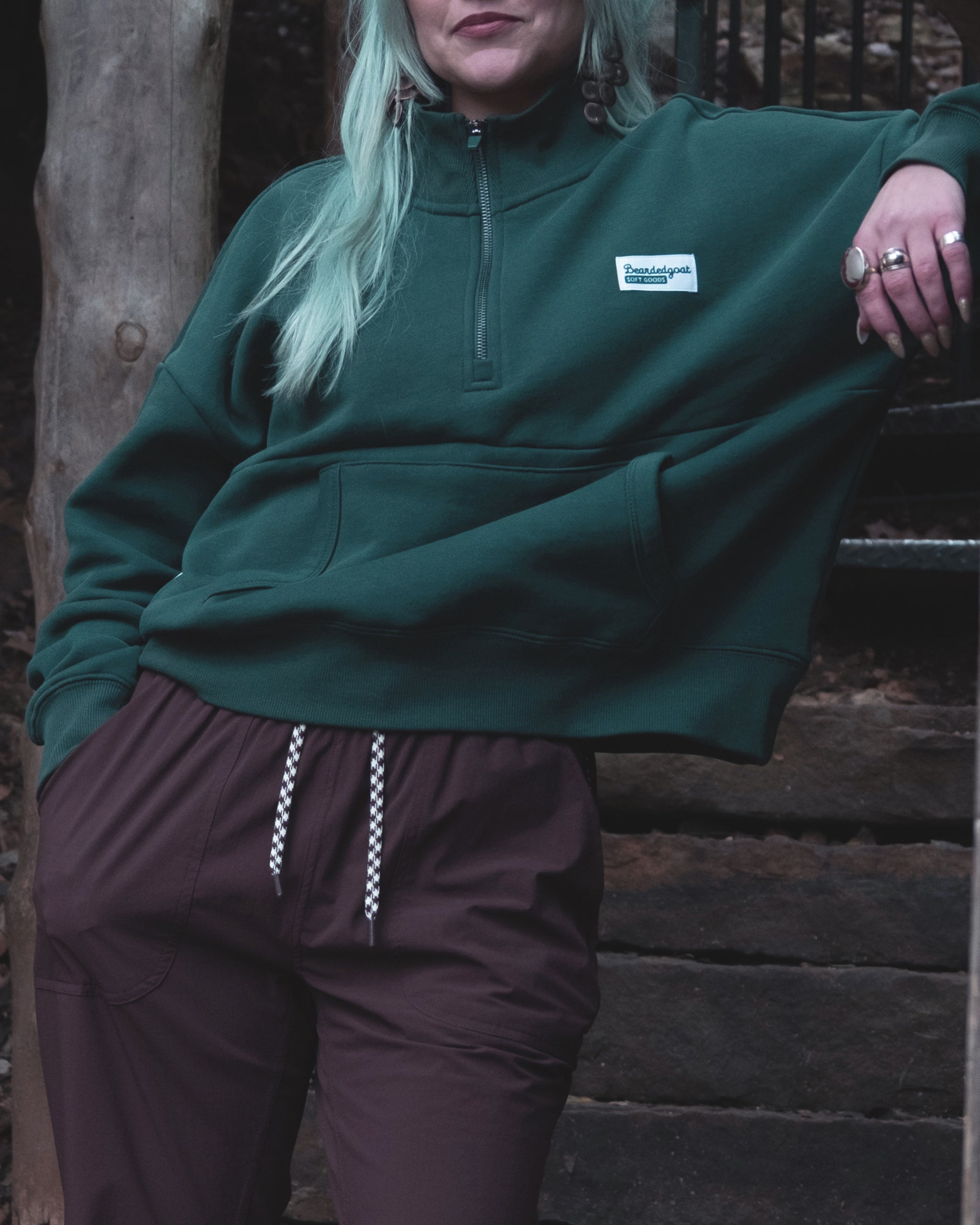 A person with light teal hair wears the BEARDEDGOAT NEW — Overnight™ Half Zip, a dark green premium cotton fleece sweatshirt, paired with brown drawstring pants. They stand outdoors, one hand in their pocket, face partially out of frame.