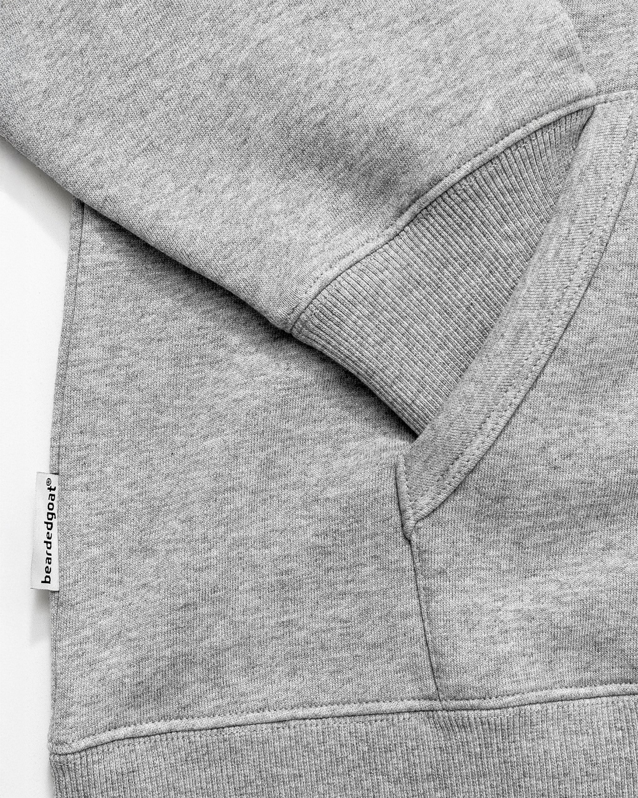 Close-up of the BEARDEDGOAT NEW — Overnight™ Half Zip sweatshirt in light gray, made from premium cotton fleece, featuring a ribbed cuff, kangaroo pocket, and a small white seam tag with "bearded edge ape" text.