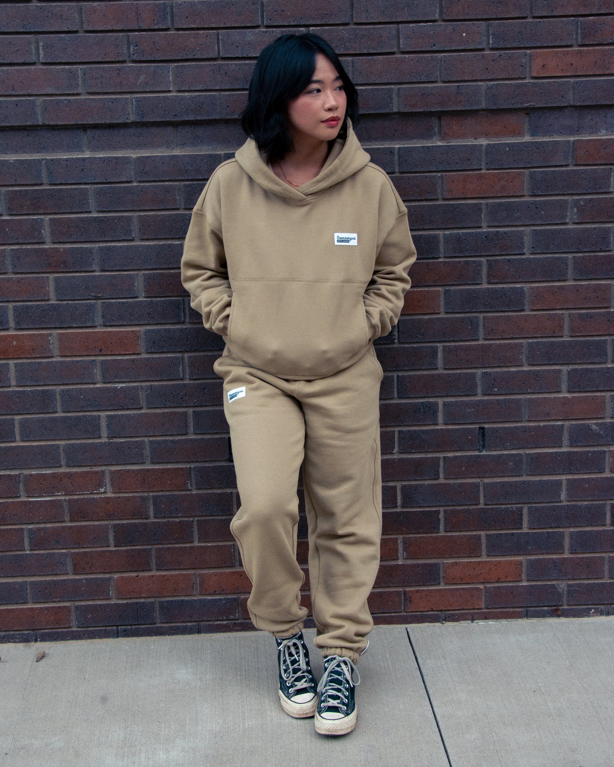A person with short, dark hair wears a tan hoodie and BEARDEDGOAT Women’s Overnight™ Classic Jogger sweatpants with a high-rise waistband, standing against a brown brick wall with hands in pockets, looking to the side in black patterned sneakers.