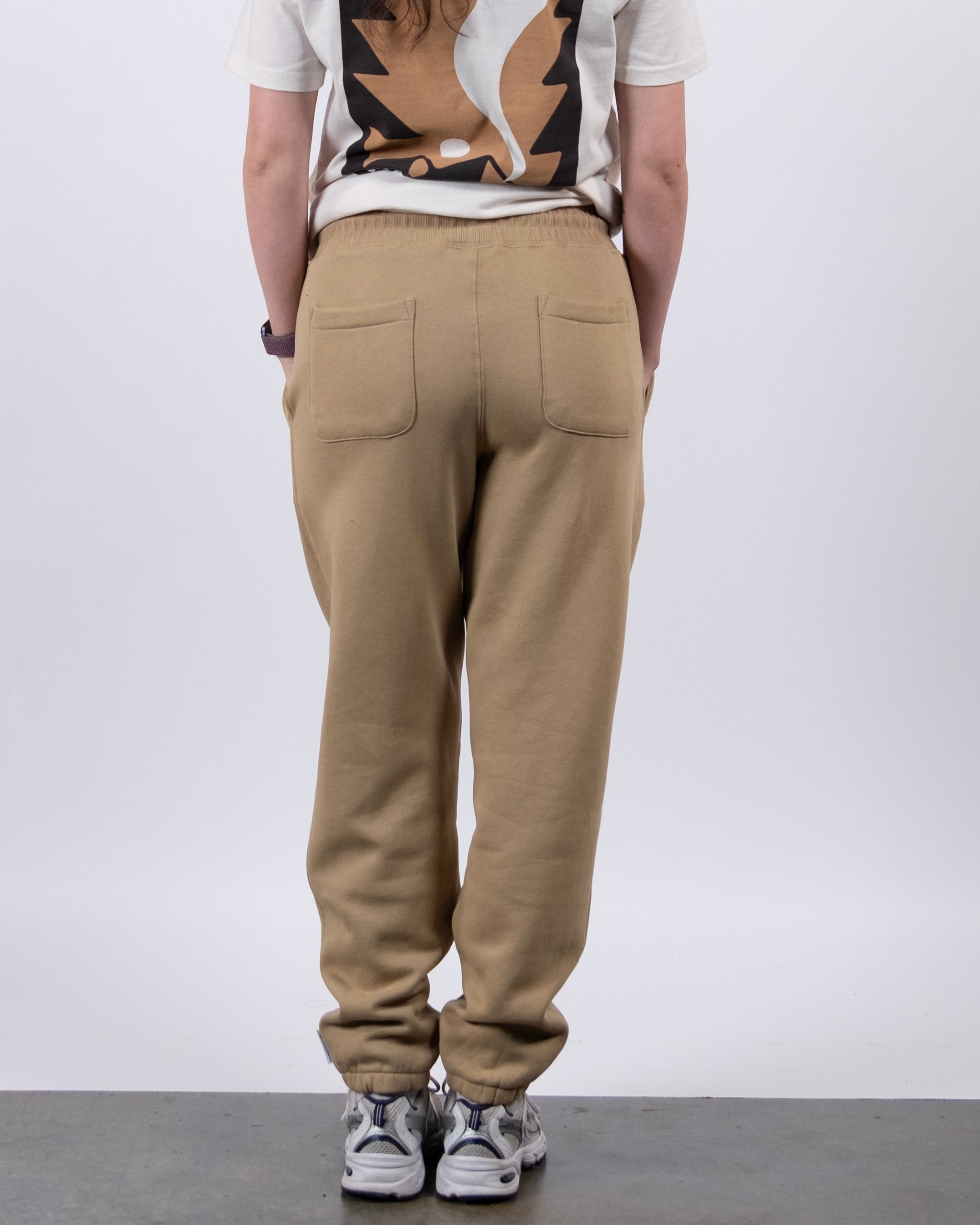 A person faces away from the camera wearing BEARDEDGOAT's NEW — Overnight™ Classic Jogger in light brown, paired with white sneakers and a bold graphic white t-shirt. The plain, light-colored background highlights the outfit.