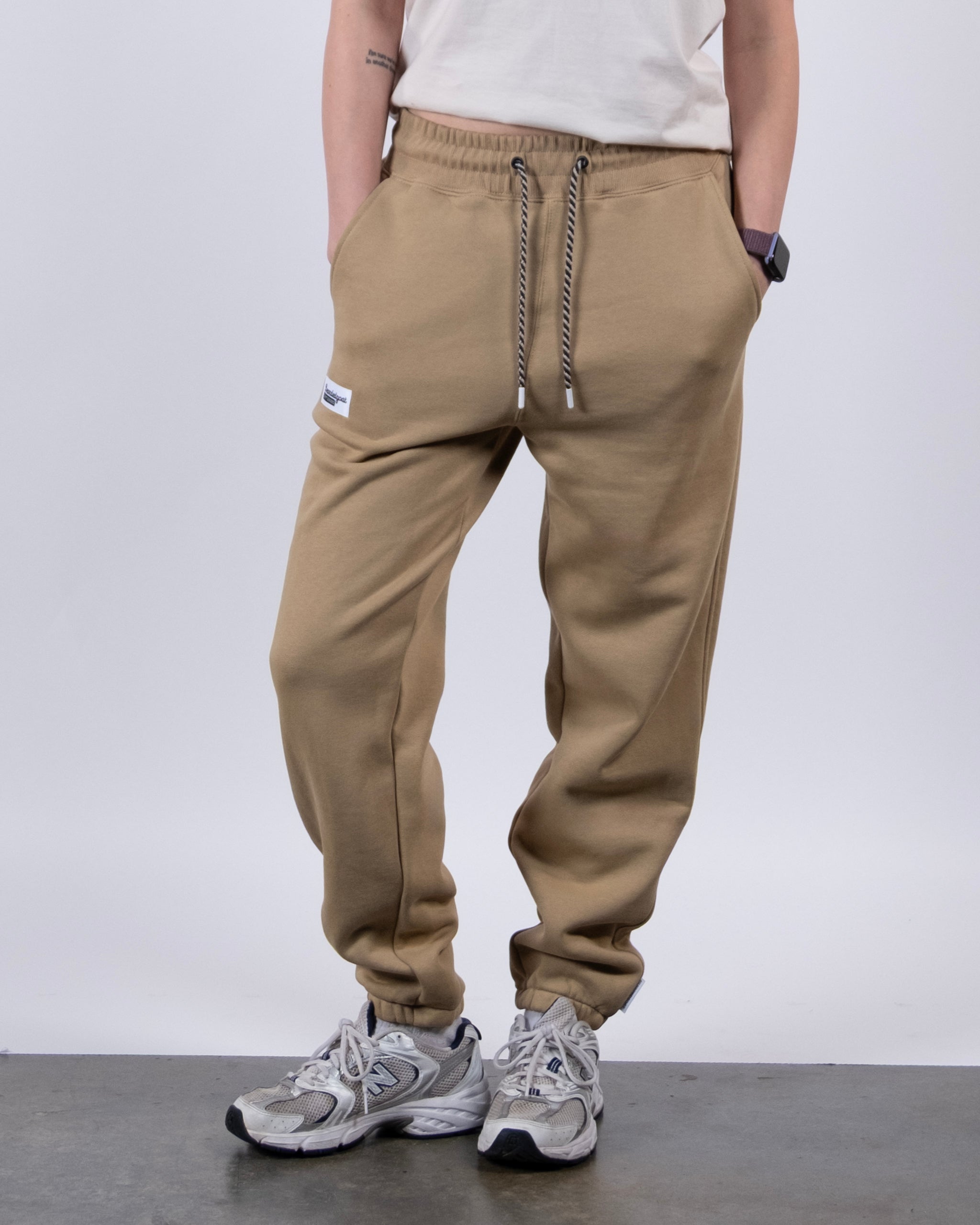 Model wears BEARDEDGOAT's NEW — Overnight™ Classic Jogger in tan, paired with a white tee, gray athletic shoes, and a smartwatch. Standing hands-in-pockets against a plain light background; face not visible.