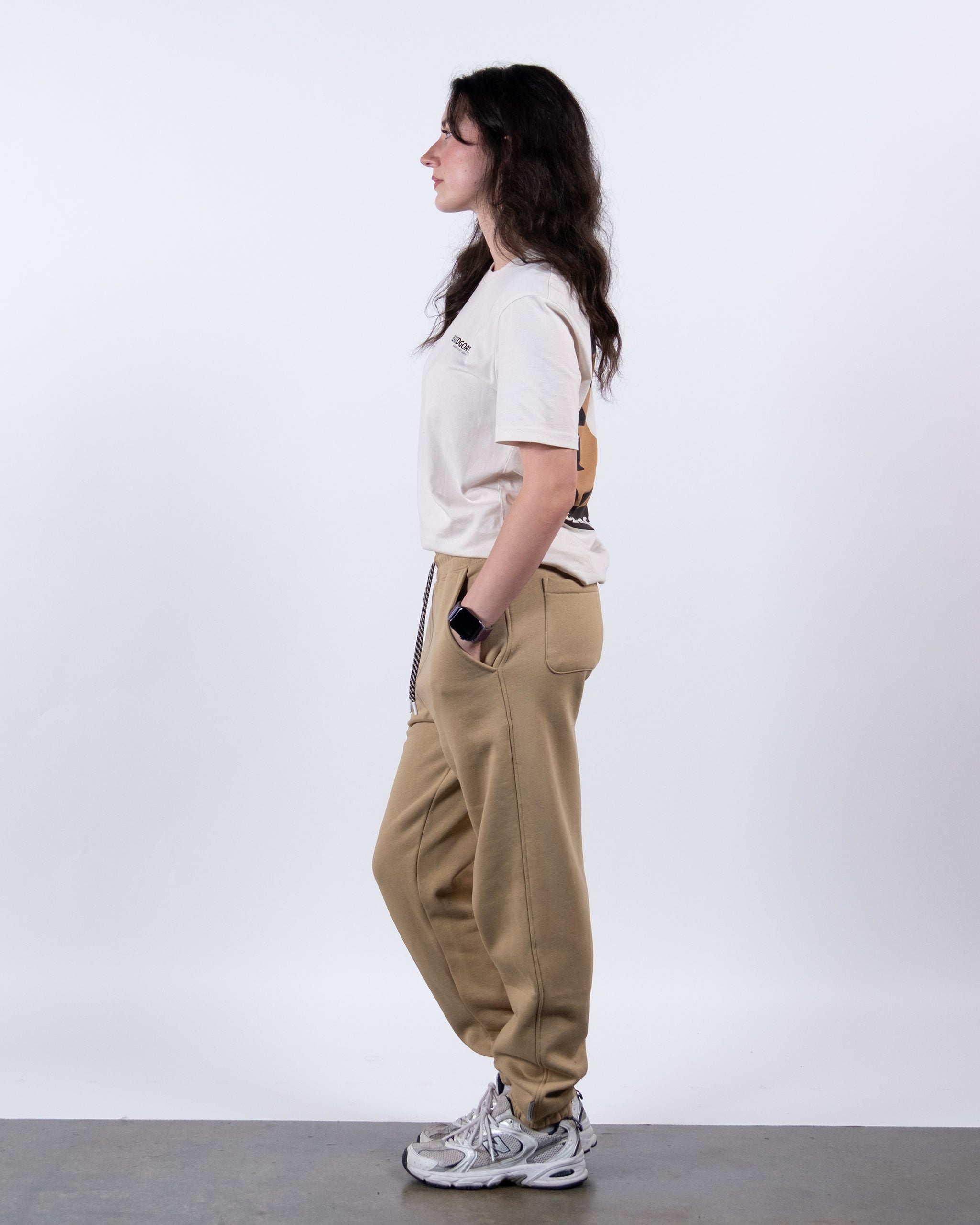 A person with long brown hair stands in profile against a white background, wearing a white t-shirt, BEARDEDGOAT NEW — Overnight™ Classic Jogger tan sweatpants, white sneakers, and a smartwatch.