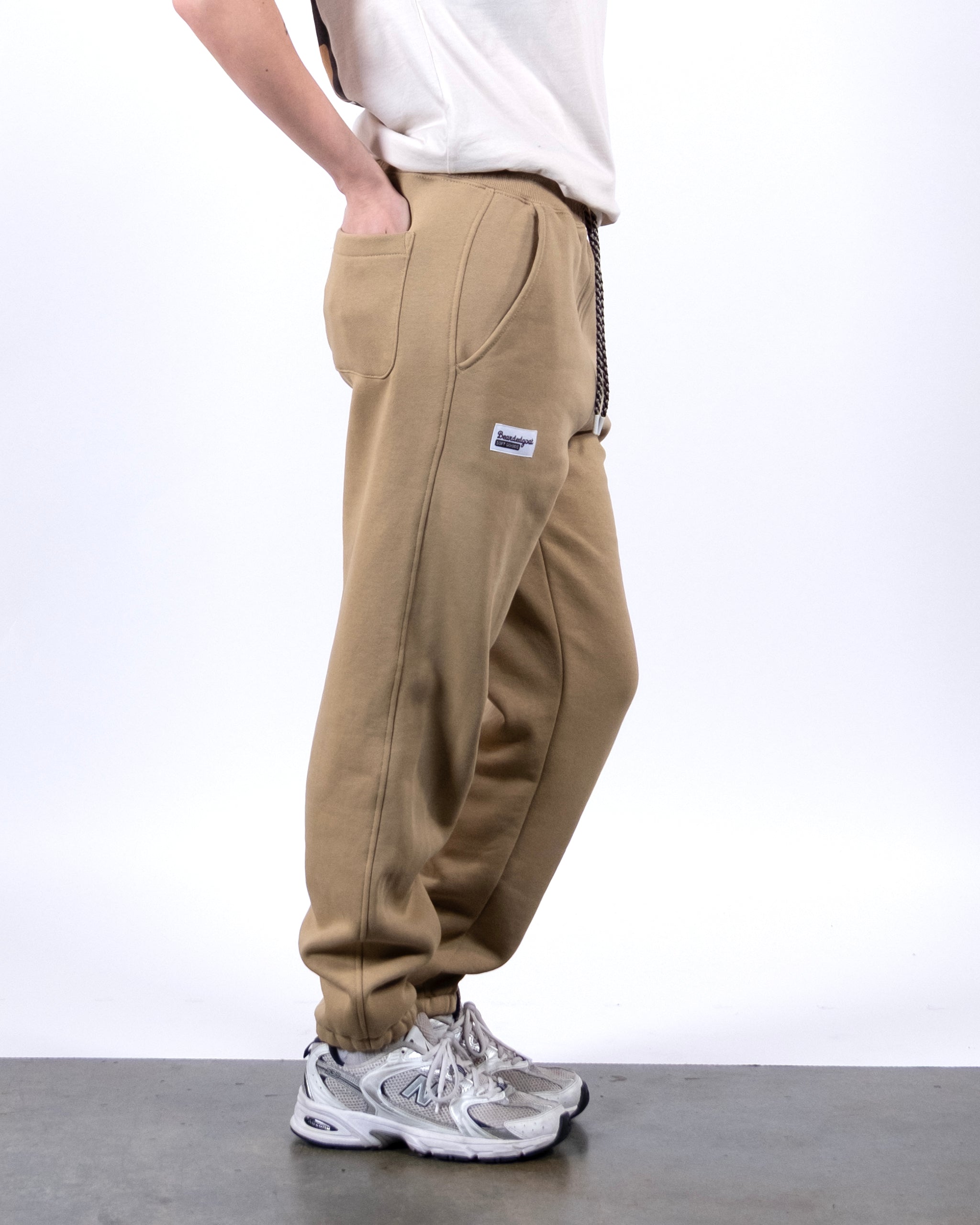 A person stands sideways in BEARDEDGOAT's NEW — Overnight™ Classic Jogger, featuring a high-rise waistband and small rectangular thigh patch. They wear a white t-shirt and silver athletic shoes, set against a plain white background.