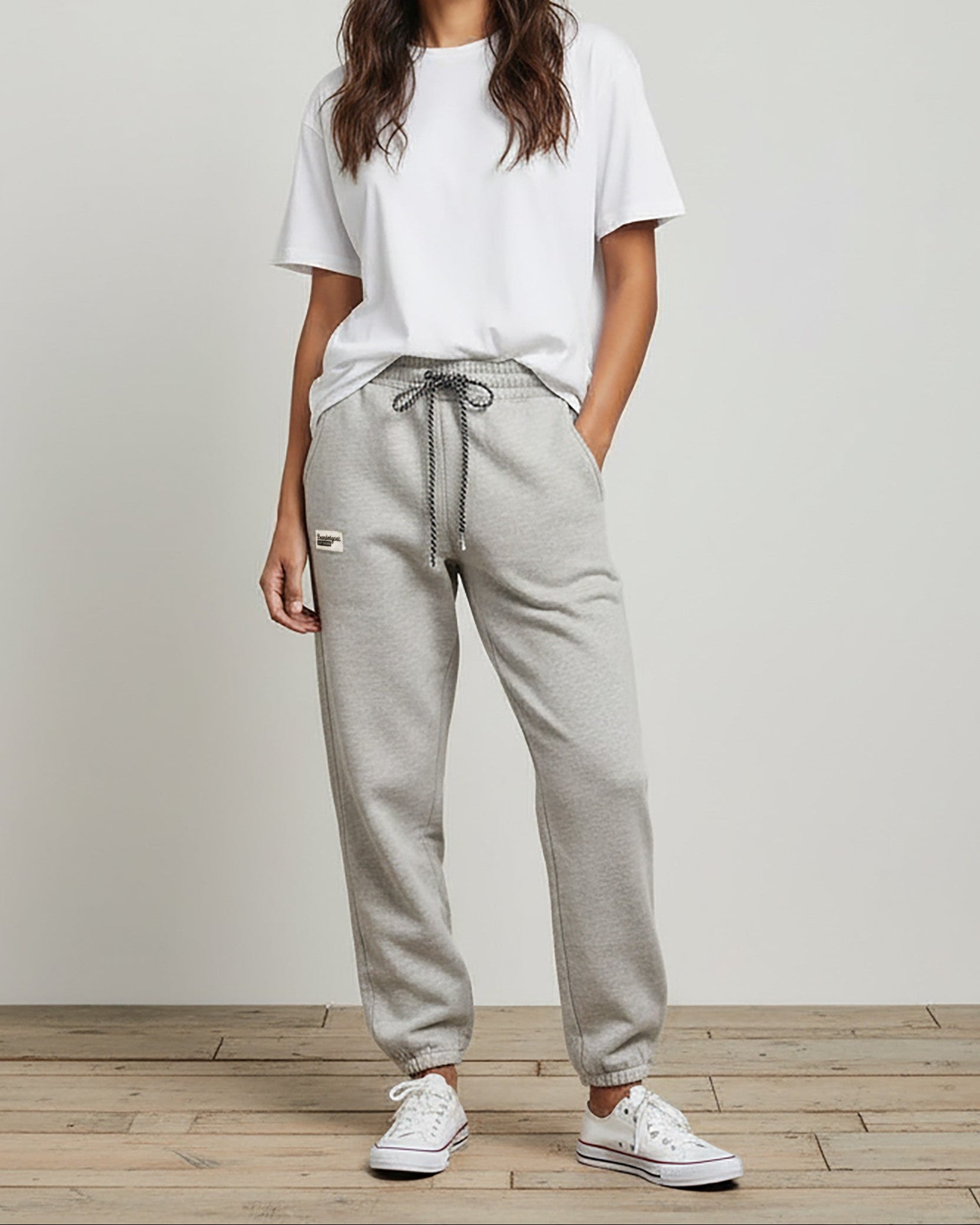 A woman stands indoors against a plain light background, hands in her pockets, wearing the BEARDEDGOAT Women’s Overnight™ Classic Jogger with a high-rise waistband, a white T-shirt, white sneakers, and her long wavy hair down.