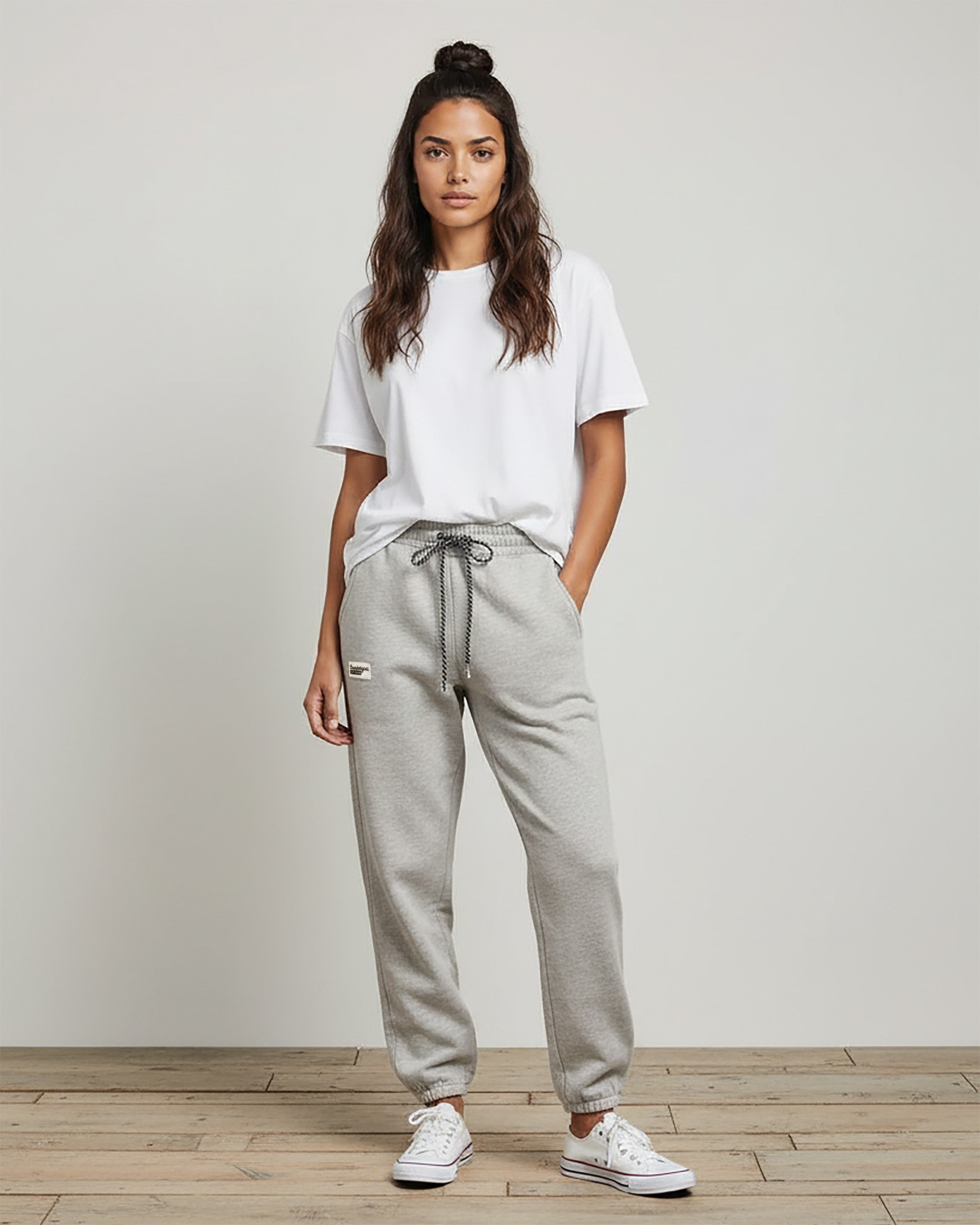 A woman stands indoors against a plain light background, hands in her pockets, wearing the BEARDEDGOAT Women’s Overnight™ Classic Jogger with a high-rise waistband, a white T-shirt, white sneakers, and her long wavy hair down.